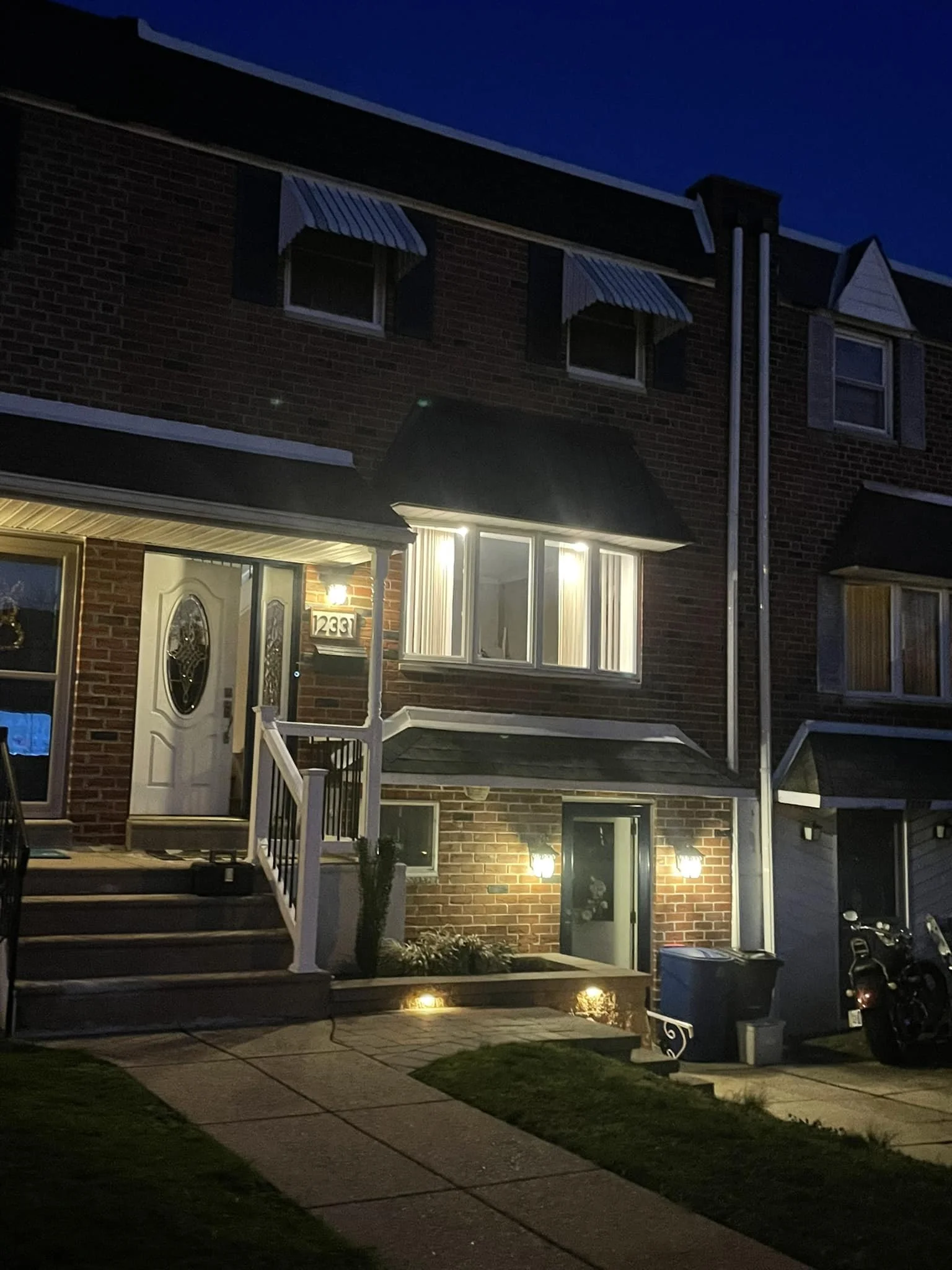 Nighttime view of a multi-story brick house with illuminated windows, front porch, and stairs leading to the front door. The house has awnings over the windows, and the yard is lit with outdoor lights.