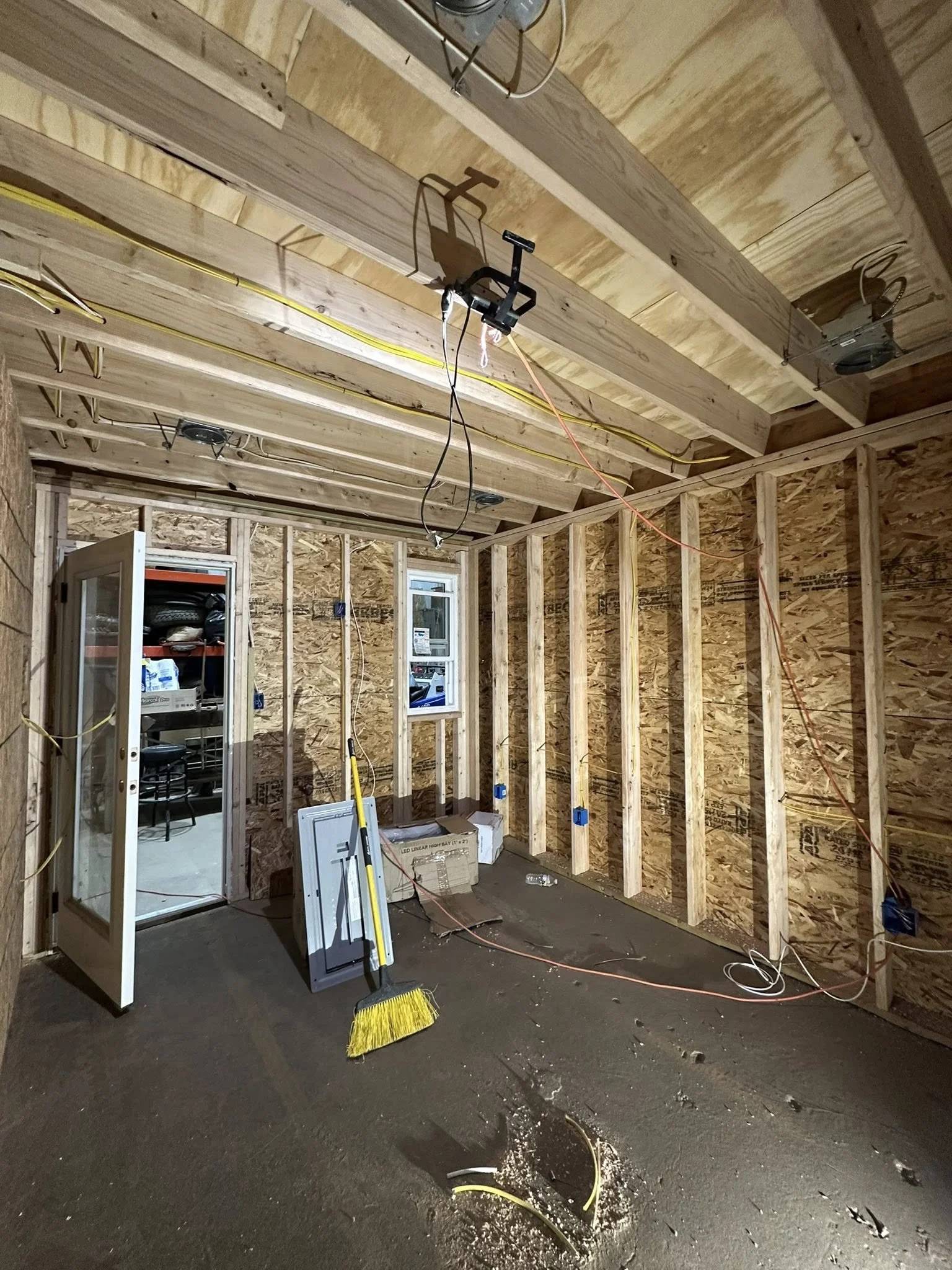 Interior of a building under construction with exposed wooden framing, electrical wiring, and insulation. Construction tools and materials are scattered on the floor, including a yellow broom, a panel, and boxes.