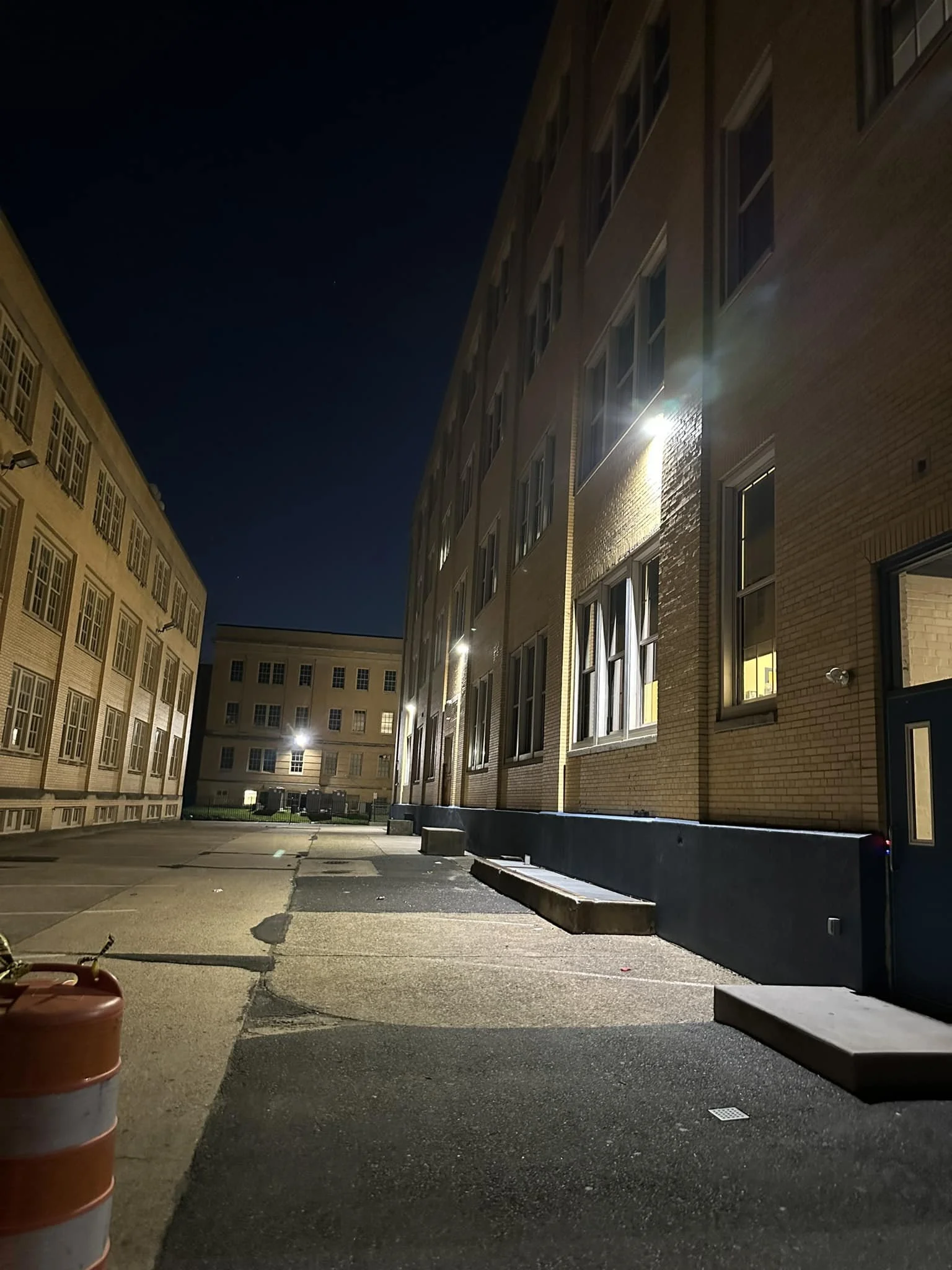 Nighttime view of an alleyway between two brick apartment buildings, with illuminated windows and exterior lights, and some construction barriers on the pavement.