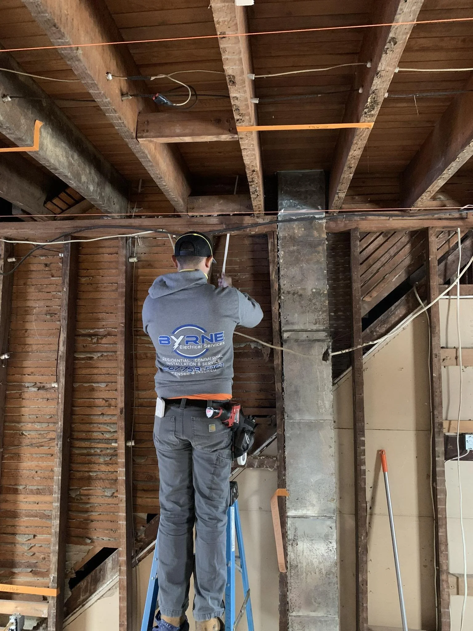 A worker on a ladder installing or repairing electrical wiring in a building with exposed wooden beams and brick walls.