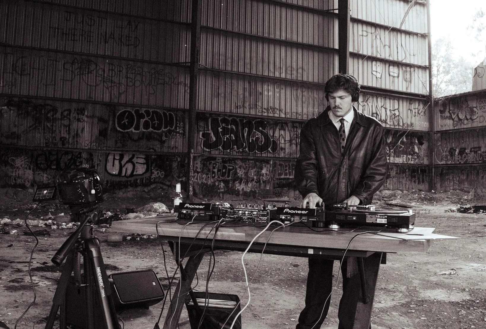 A DJ performing in an abandoned, graffiti-covered warehouse. He is wearing a leather jacket and headphones, standing behind a table with DJ equipment and a camera on a tripod nearby.