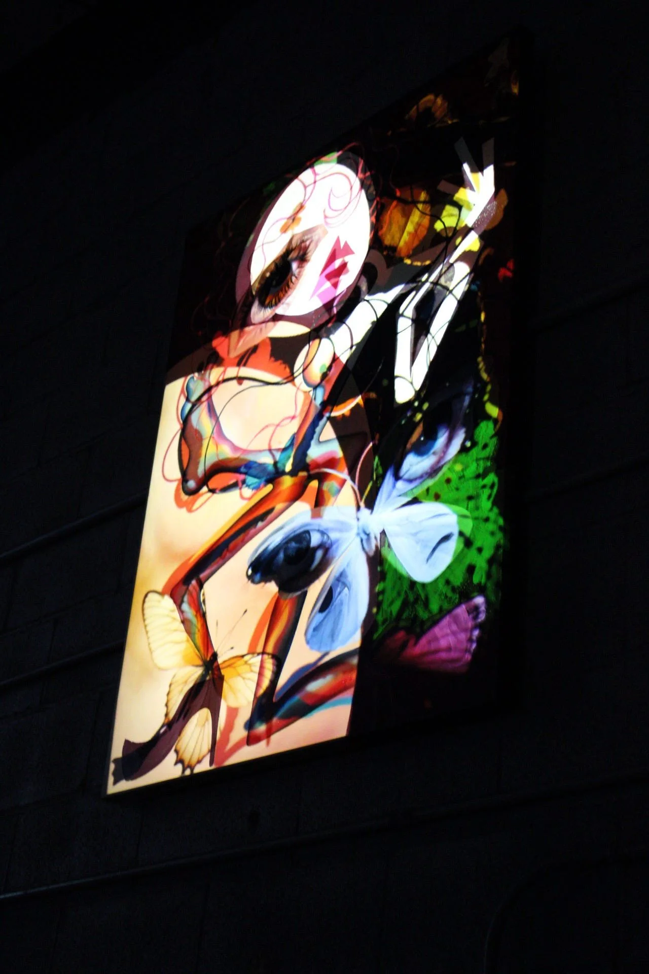 Colorful neon sign of a girl with butterfly wings, featuring large eyes and makeup, surrounded by butterflies and abstract designs on a dark background.