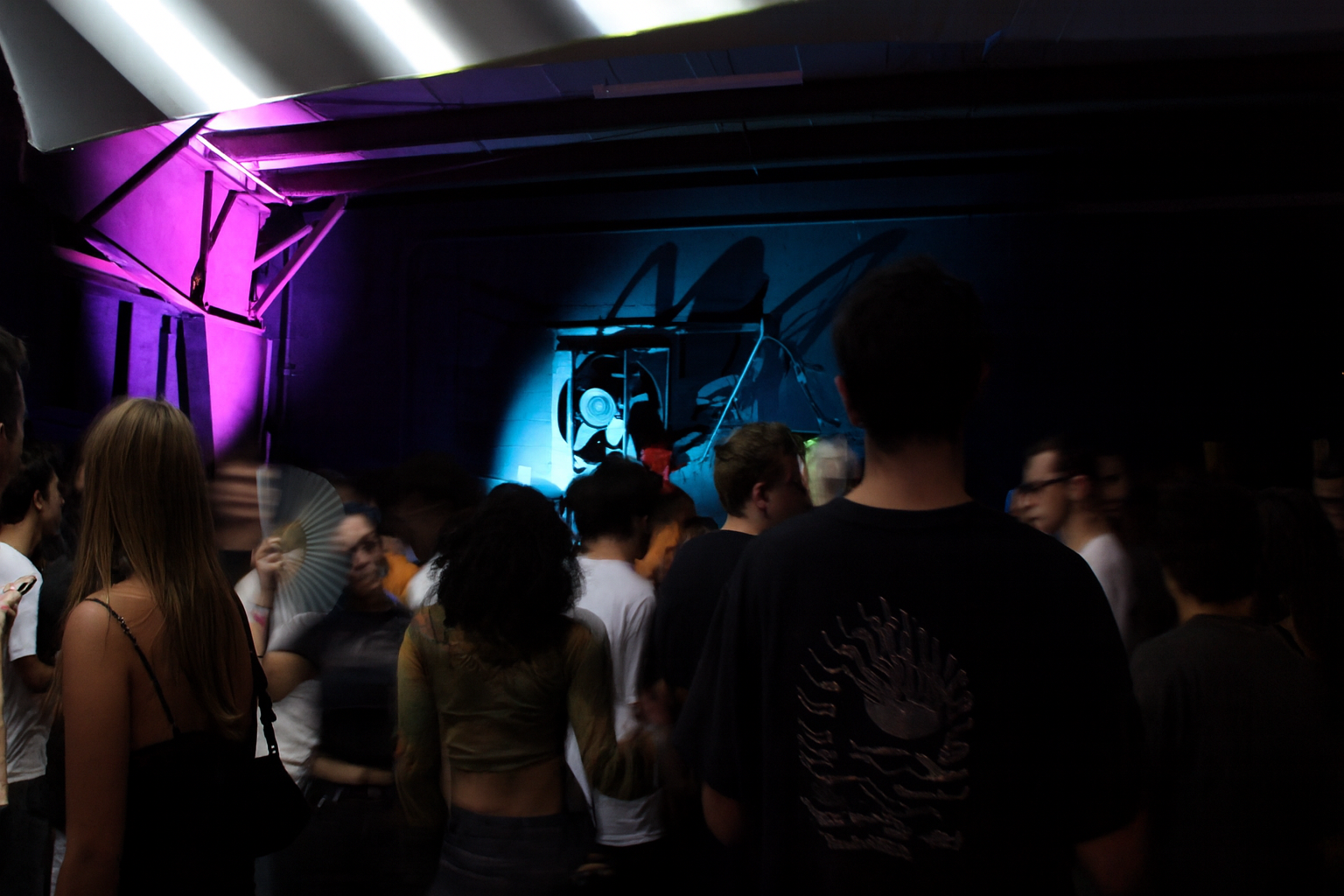 Crowd of people at an indoor event with colorful purple and blue lighting and abstract artwork on the wall.