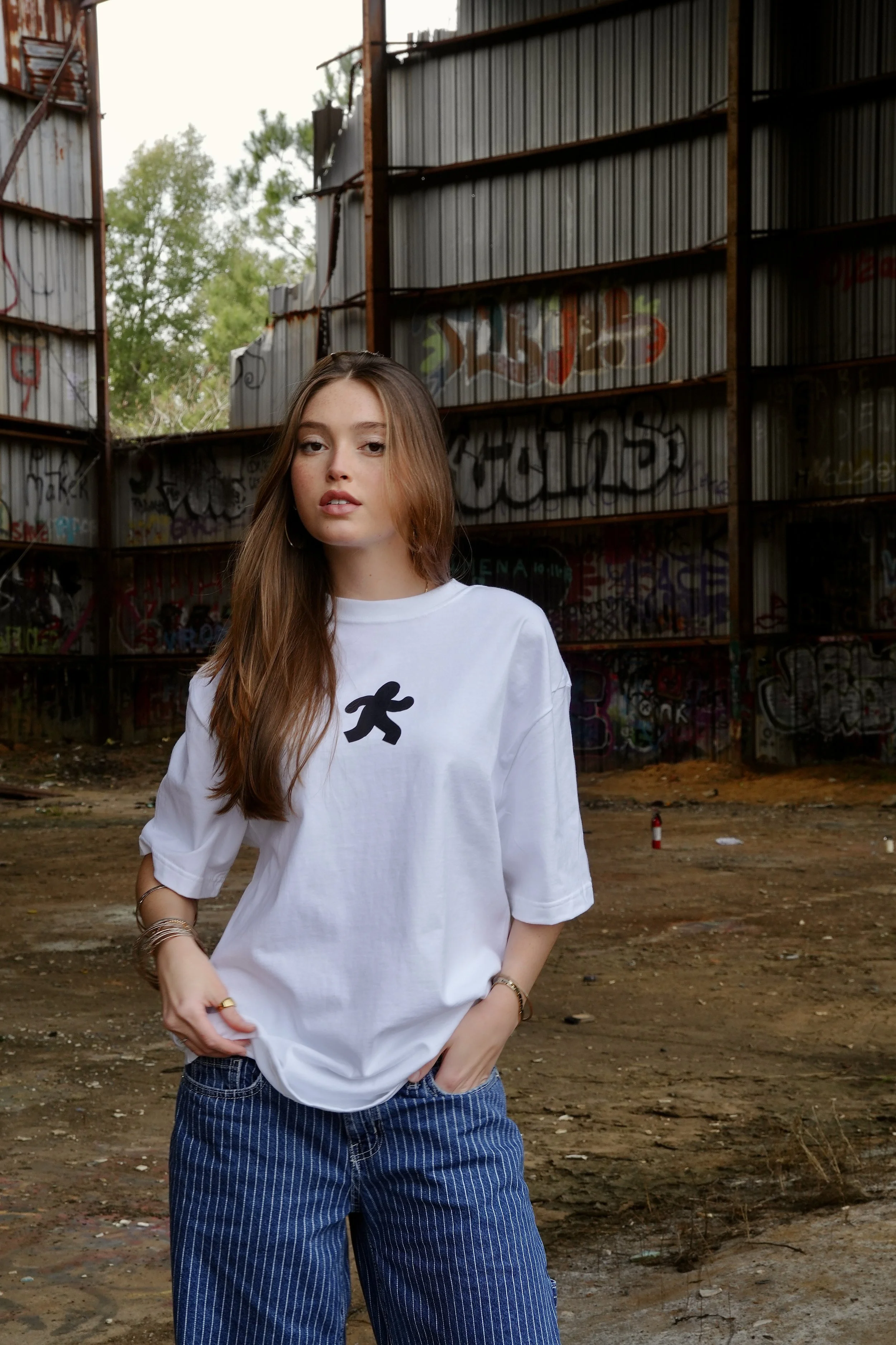 A young woman with long brown hair wearing a white T-shirt with a small graphic on the front and blue jeans, standing in front of a graffiti-covered metal structure in an outdoor, urban setting.