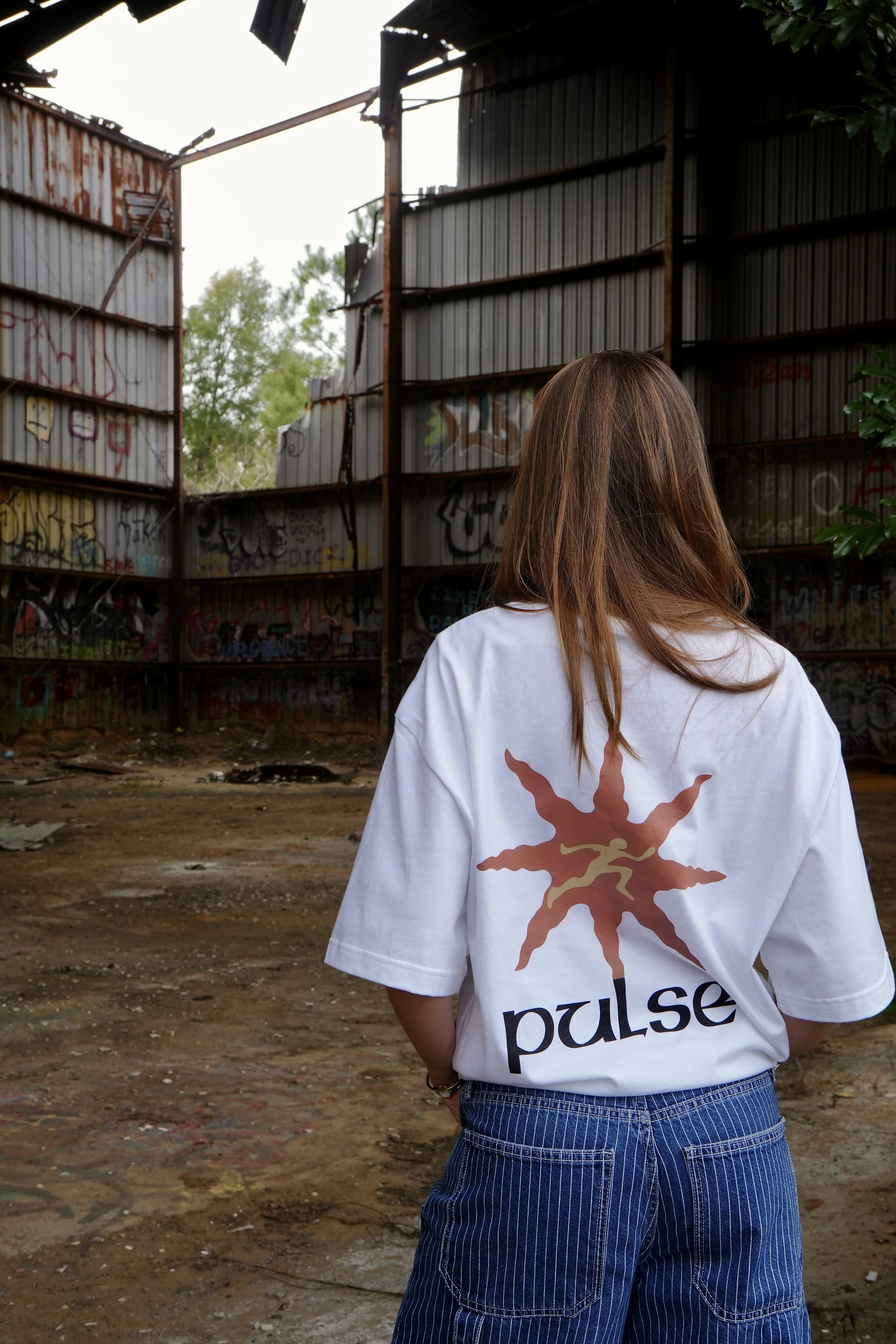 A woman with brown hair wearing a white t-shirt with a brown and tan abstract design and the word 'pulse' on the back, standing in an abandoned, graffiti-covered industrial building with an open roof and dirt ground.