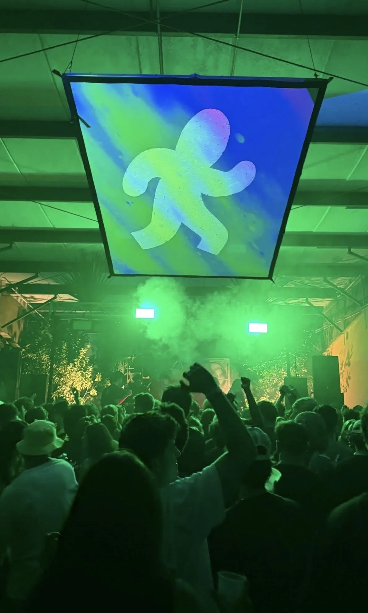 Crowd of people at an indoor music event with green lighting and a large hanging screen displaying a stylized walking man icon.