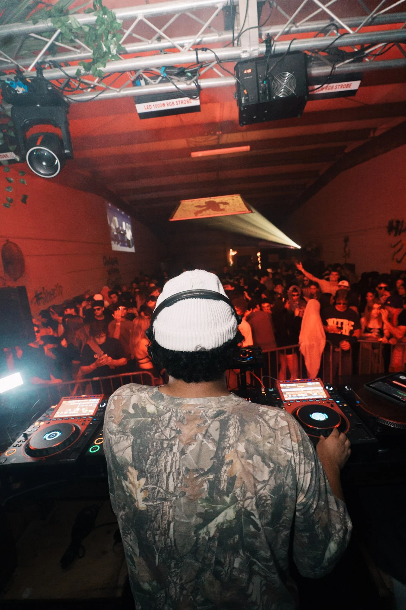 A DJ wearing a white beanie and camouflage shirt is performing at a nightclub with a crowd dancing and red lighting in the background.