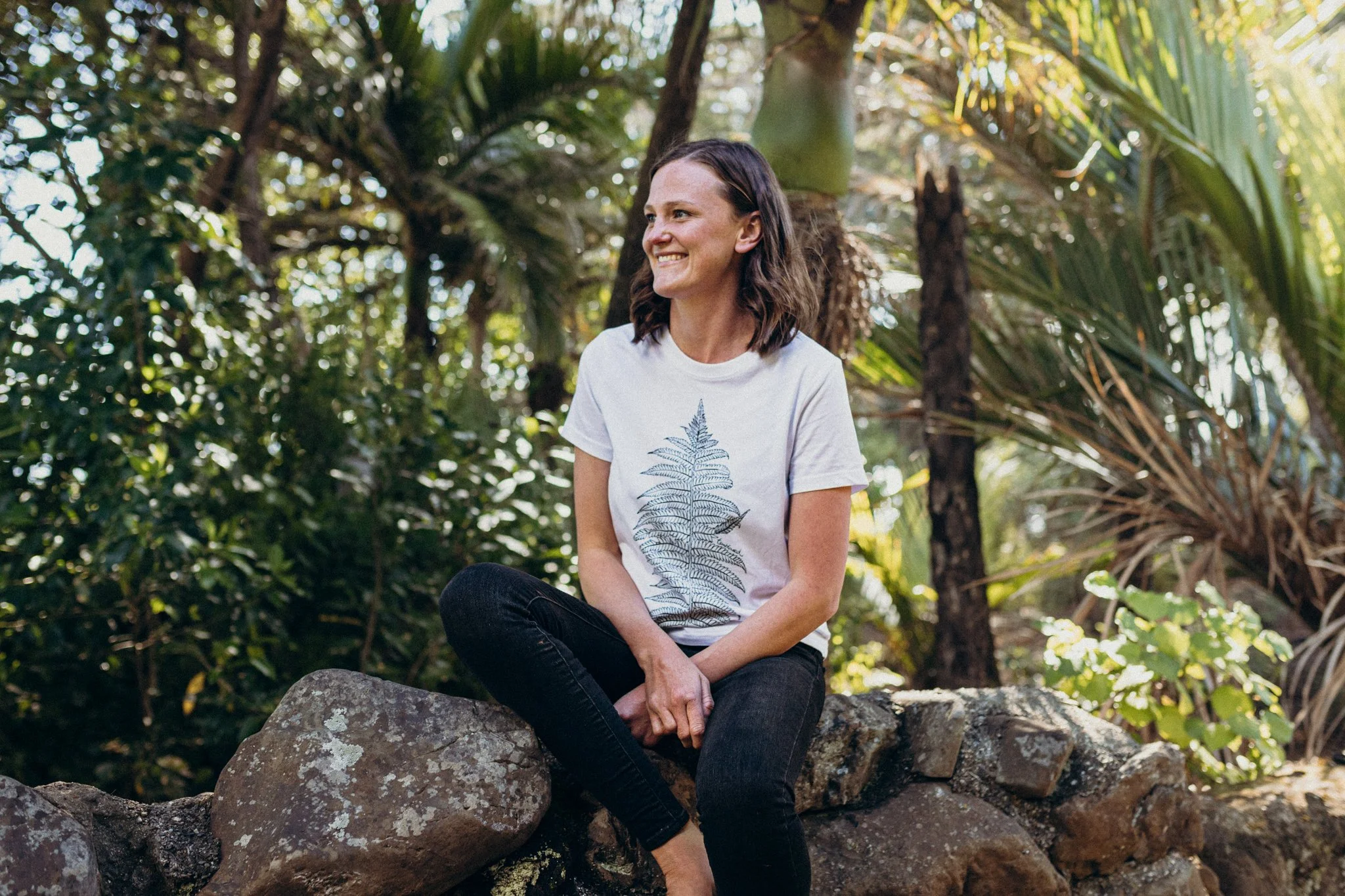 A woman sitting on a rock in a lush, green forest, smiling and looking to her left.