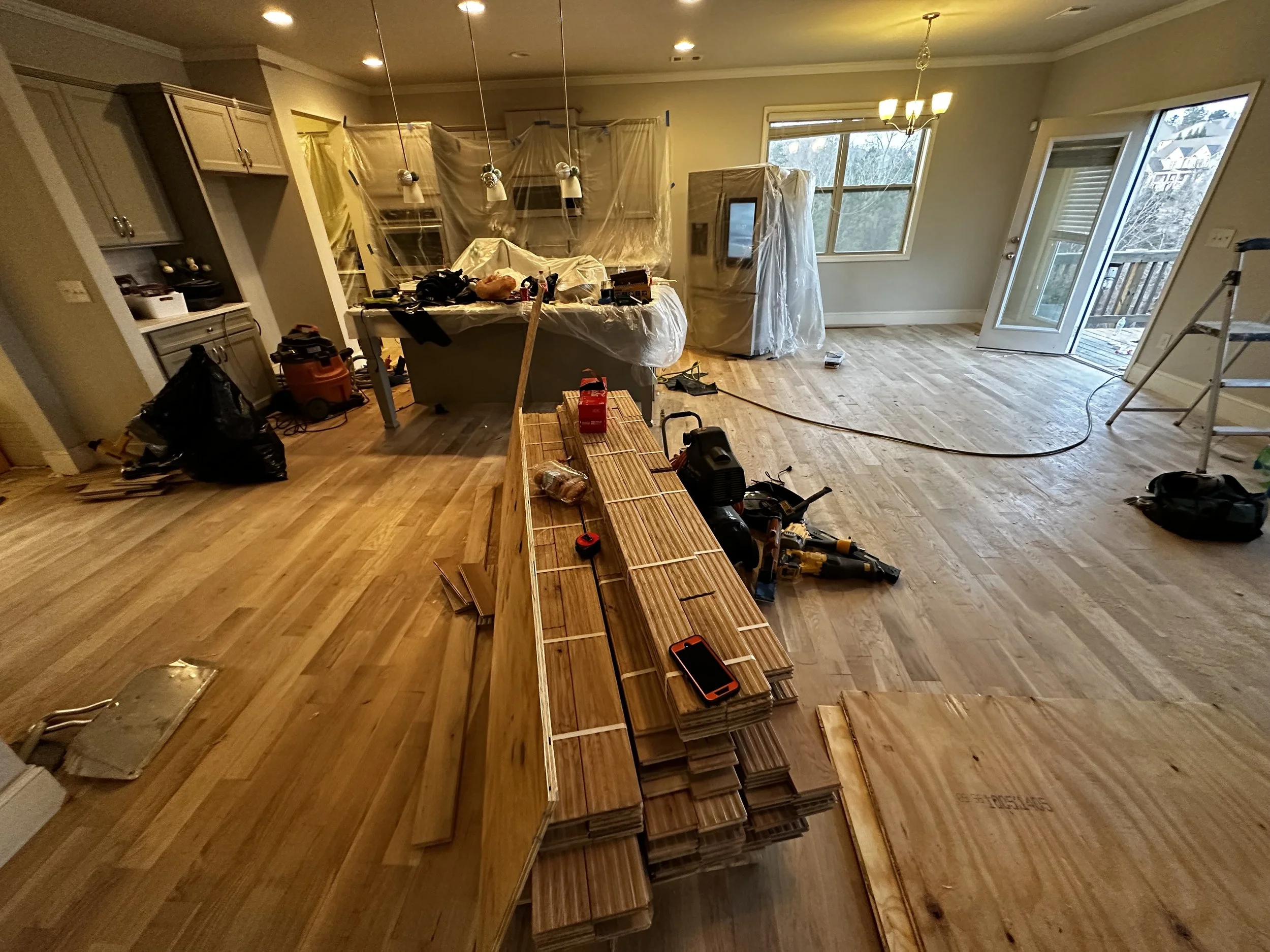 Living room during renovation with hardwood flooring being installed, tools, and furniture covered with plastic.