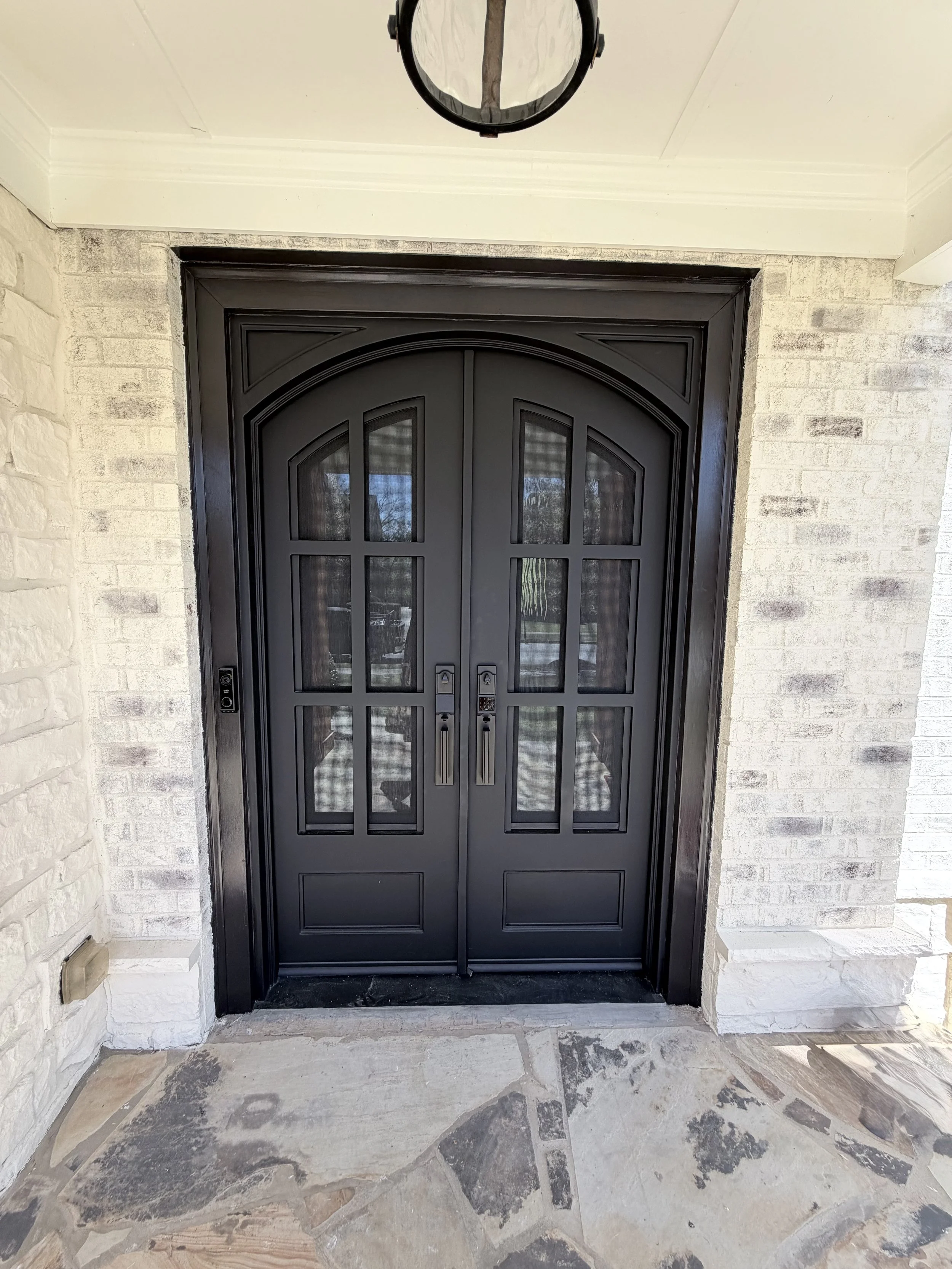 Black double front door with glass panes, surrounded by white brick walls, with a stone floor in front and a ceiling light fixture above.