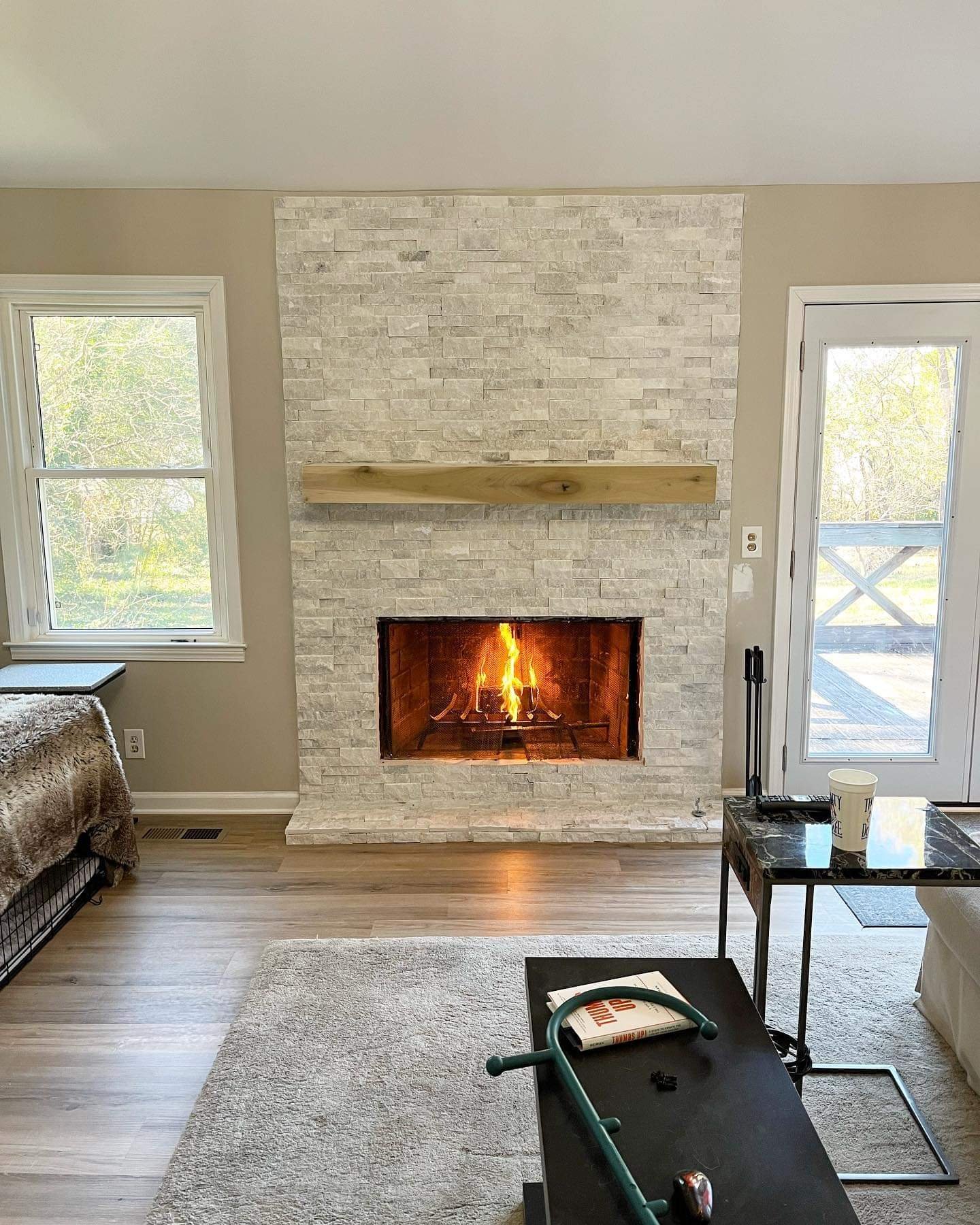 Living room with a stone fireplace and a fire burning inside, a wooden mantel above, two windows on either side, a glass door on the right, a small table with a cup, books, and a stethoscope, and a beige rug on light wood flooring.