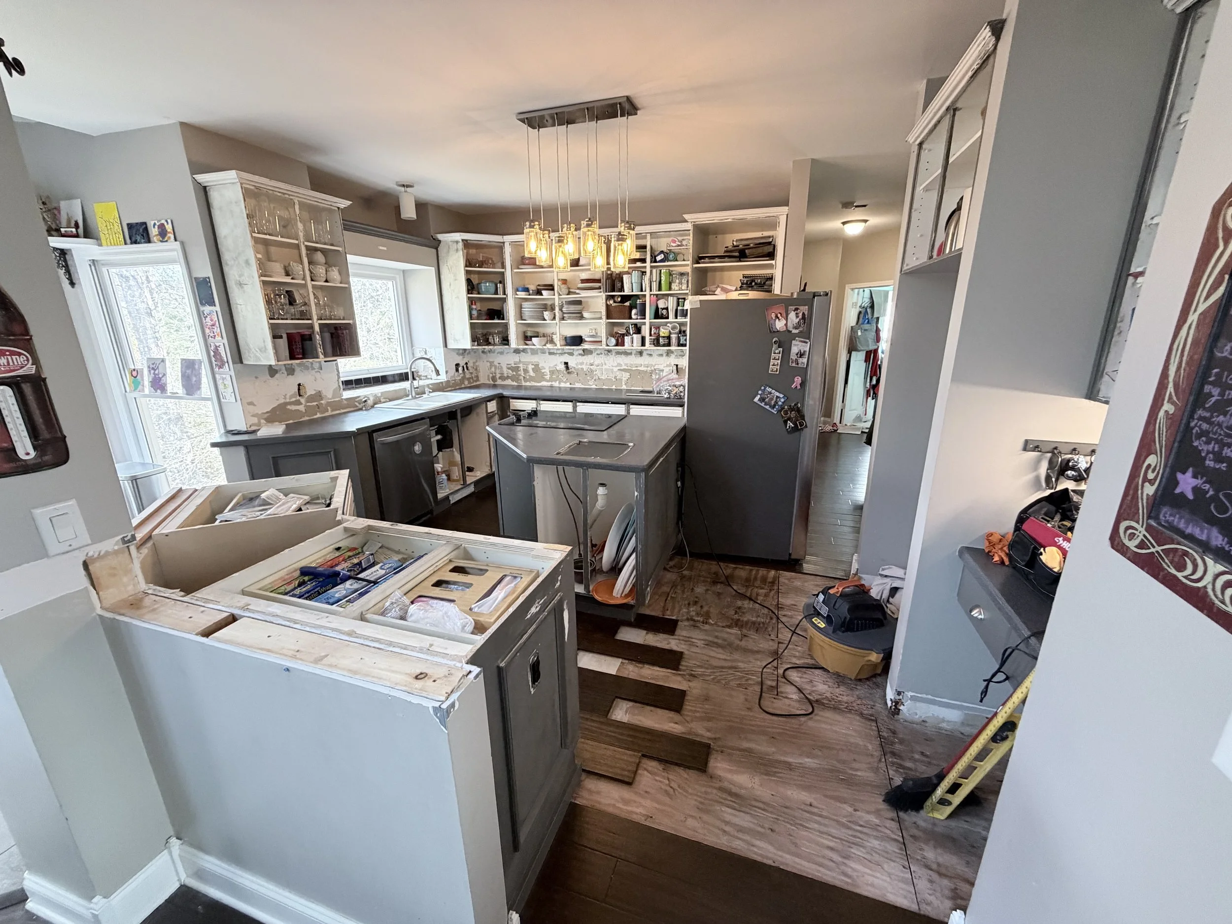 Kitchen undergoing renovation with cabinets, countertops, a refrigerator, and construction tools visible.