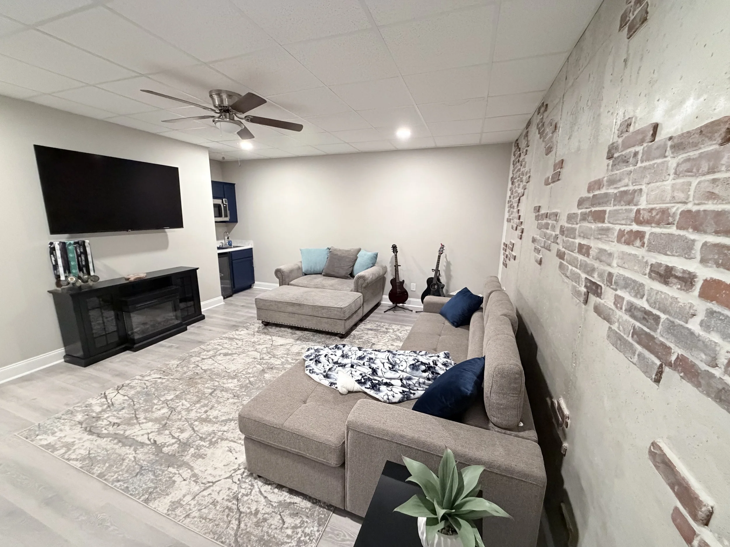 Living room with beige sectional sofa, gray armchair with ottoman, TV on black stand, brick accent wall, three guitars on stands, patterned area rug, potted plant, wall-mounted TV, small kitchenette with blue cabinets, ceiling fan, and white ceiling tiles.