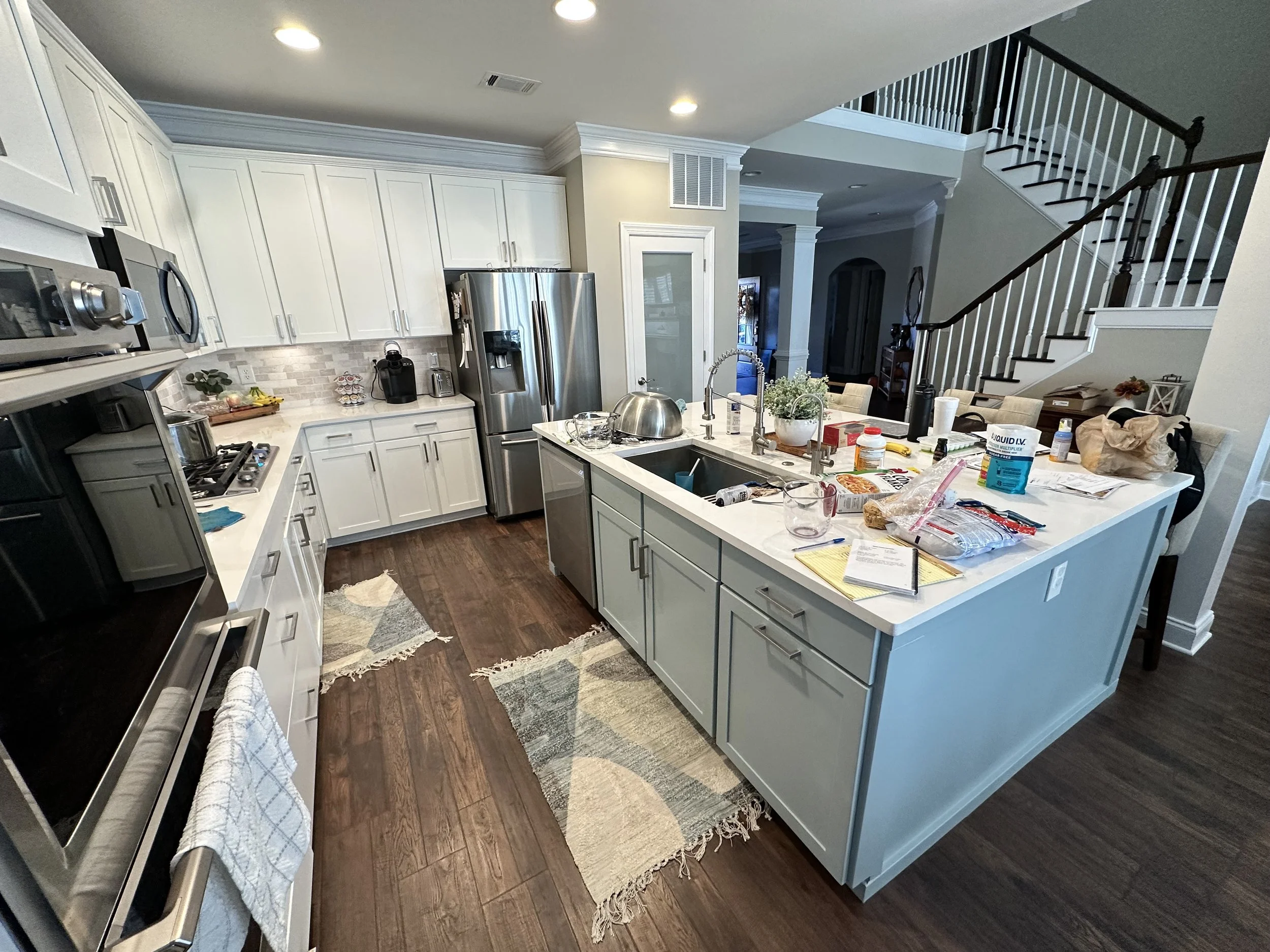 Messy kitchen with white cabinets, stainless steel appliances, and a kitchen island filled with food, papers, and various items.