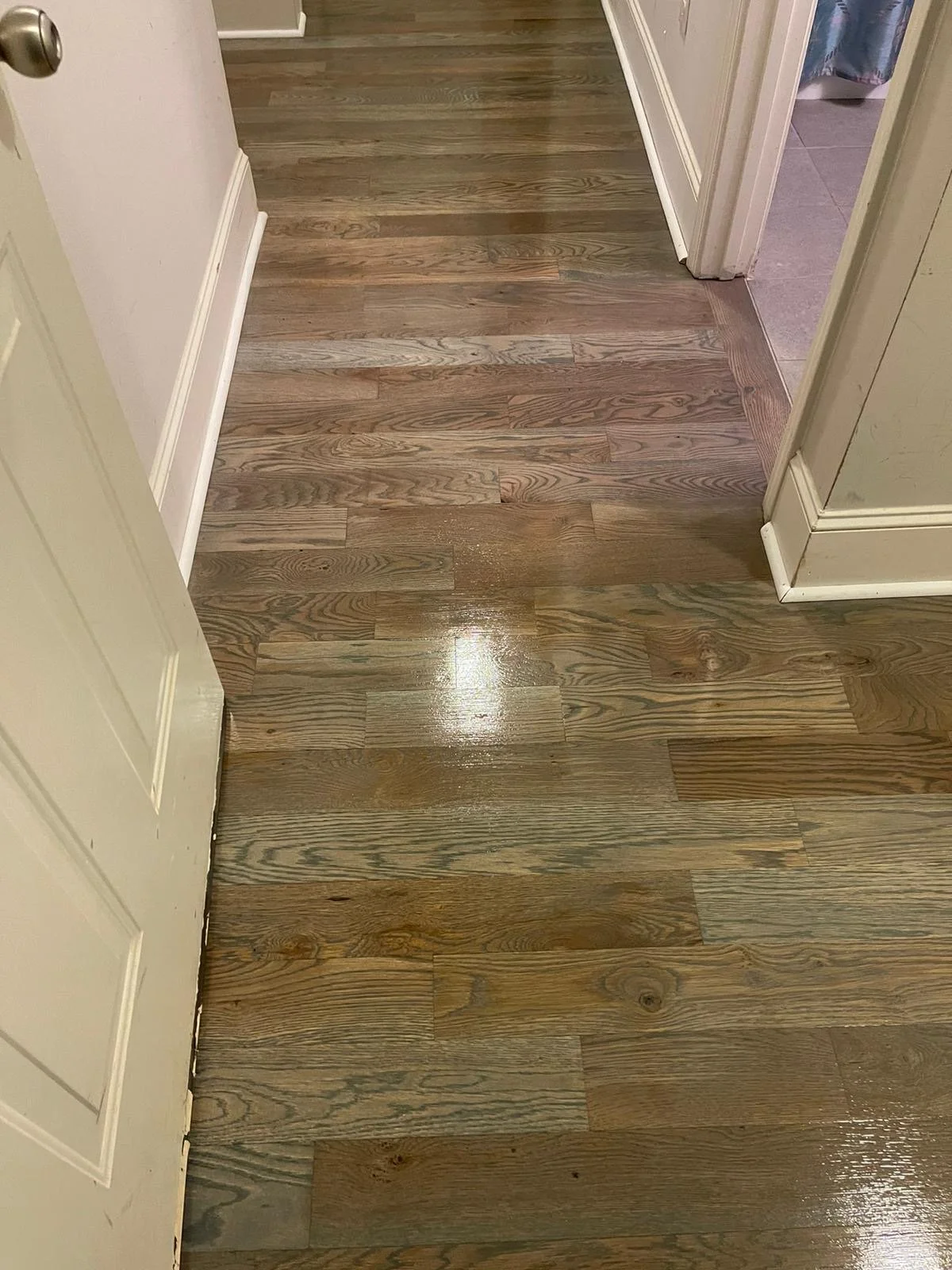 Polished wooden floor in a hallway leading to a bathroom, with bathroom tiles visible through an open door.