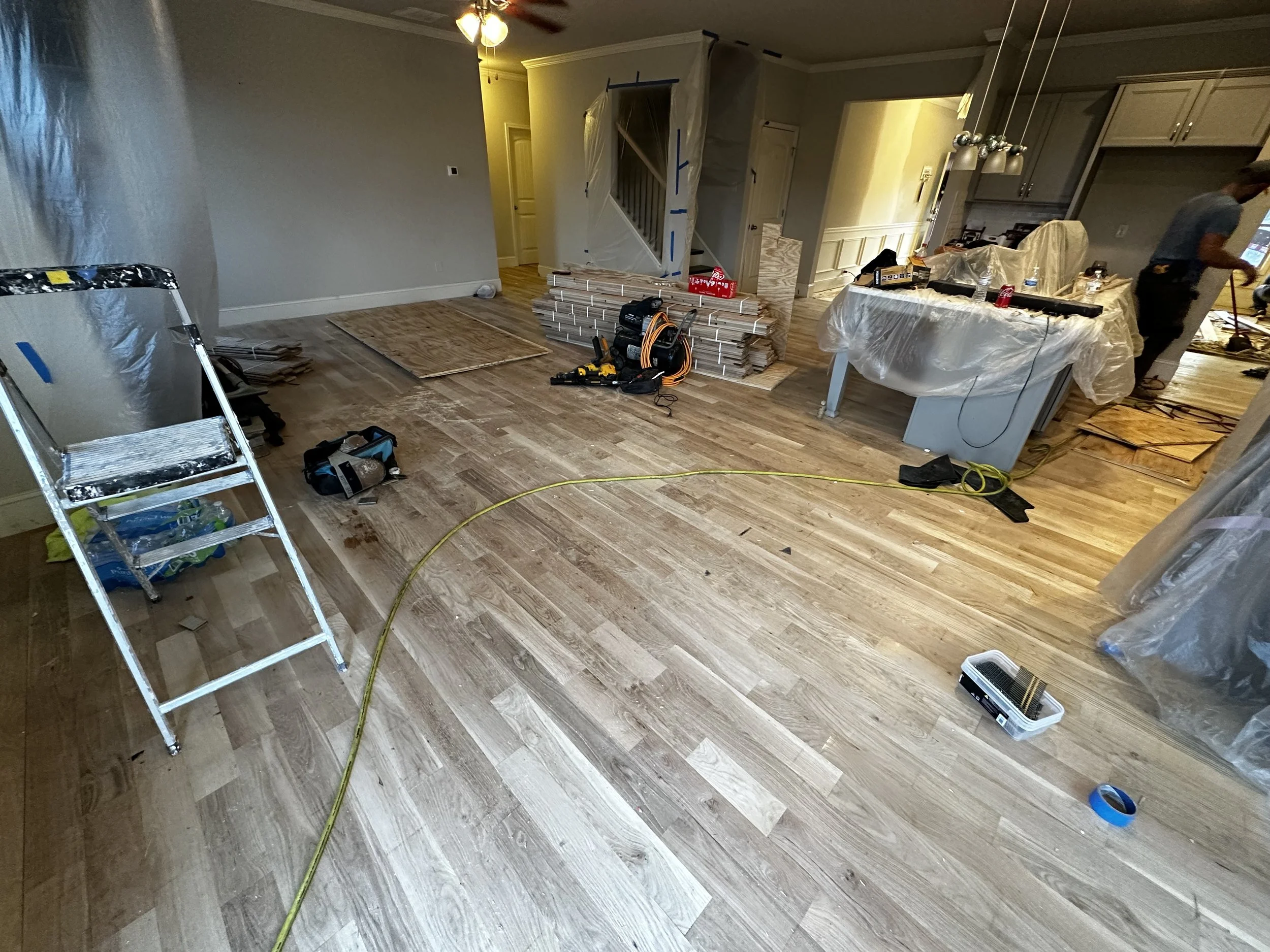 Living room undergoing hardwood floor installation with tools, materials, ladder, and workers in the background.