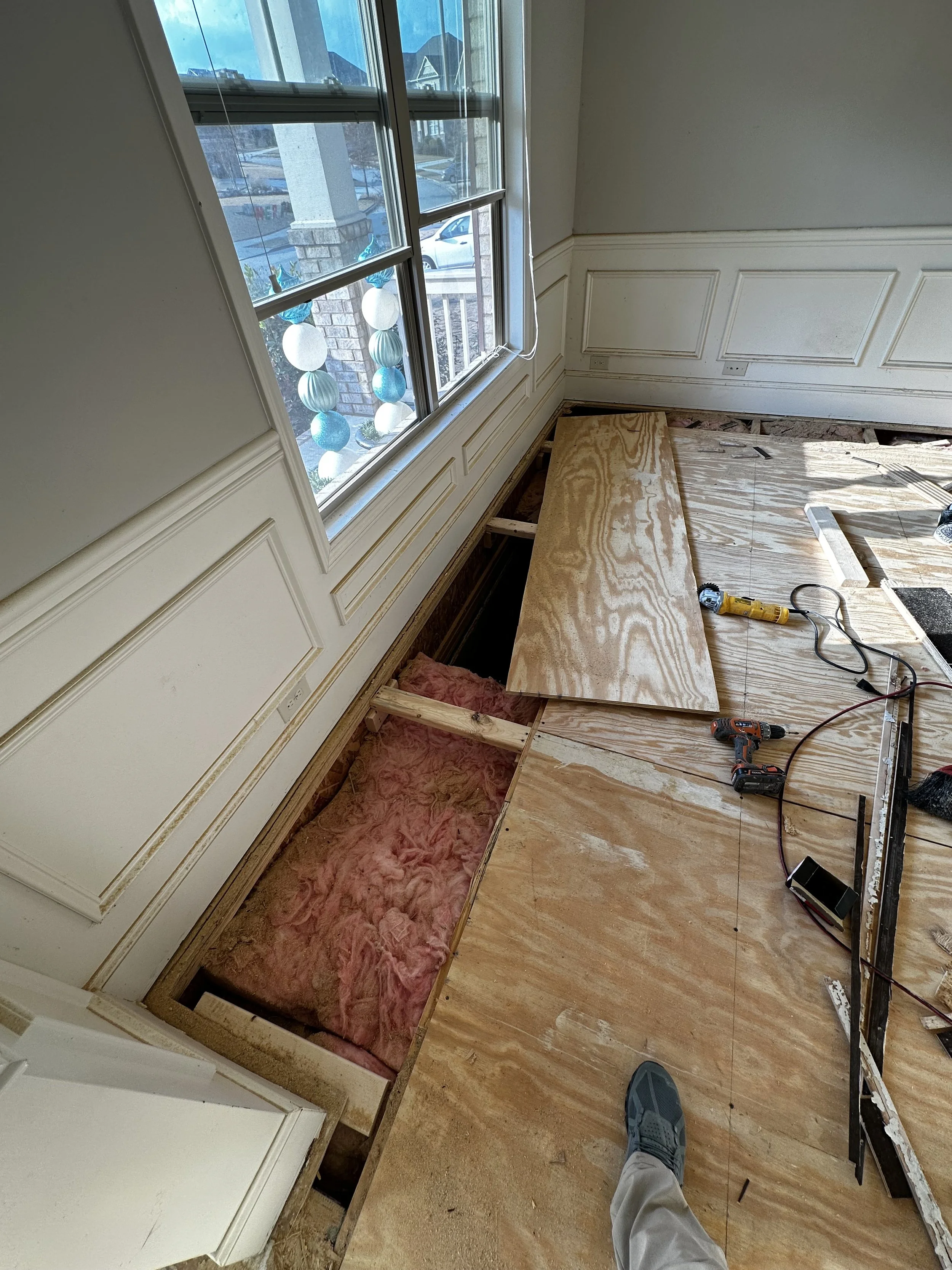 Room under construction with plywood flooring and pink insulation, tools like a drill and a saw on the floor, and a large window with decorative hanging ornaments outside.