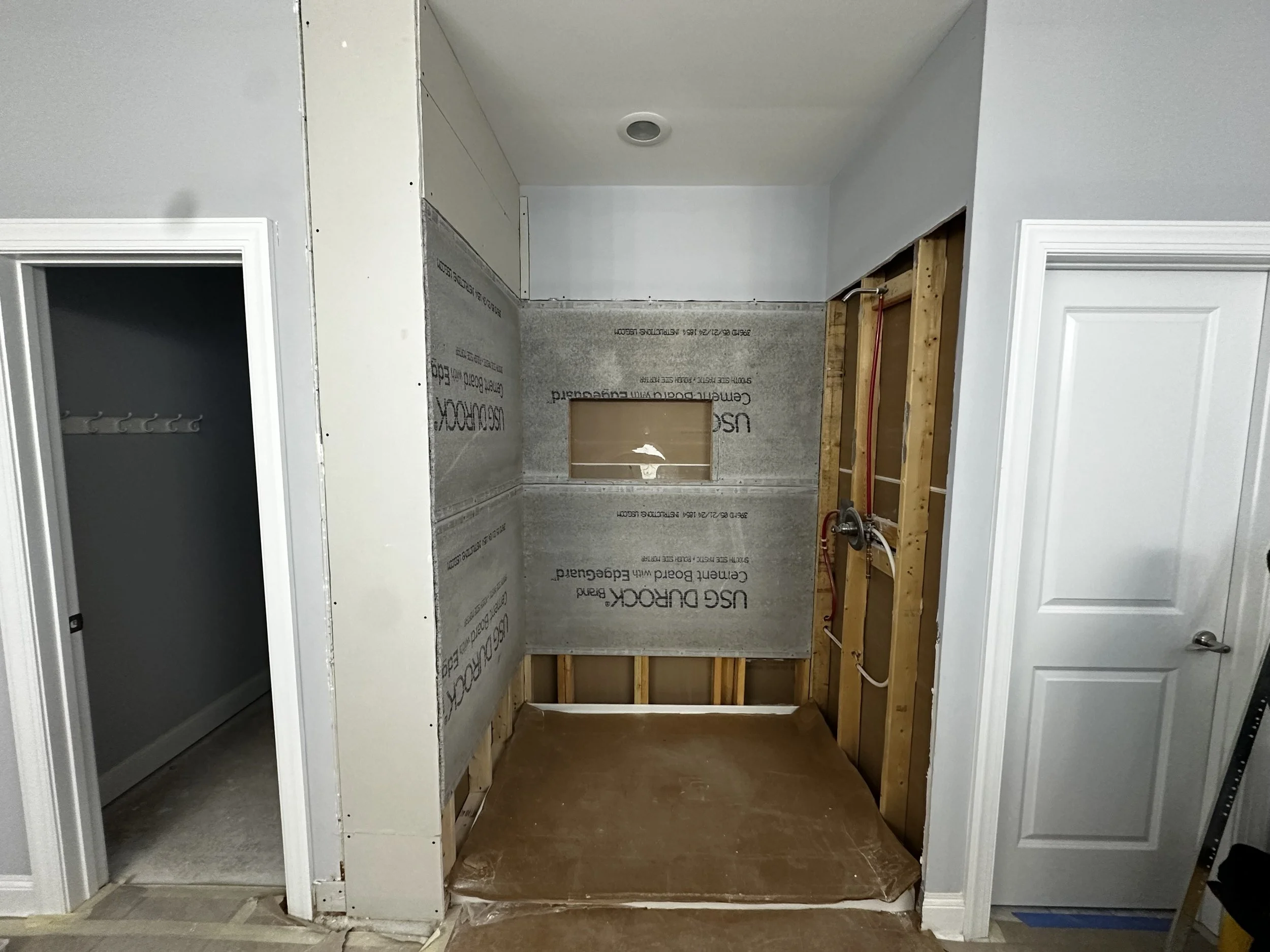 Room under renovation with exposed wall framing, insulation, and plumbing, with a small window opening and an adjacent closet on the left with hooks.