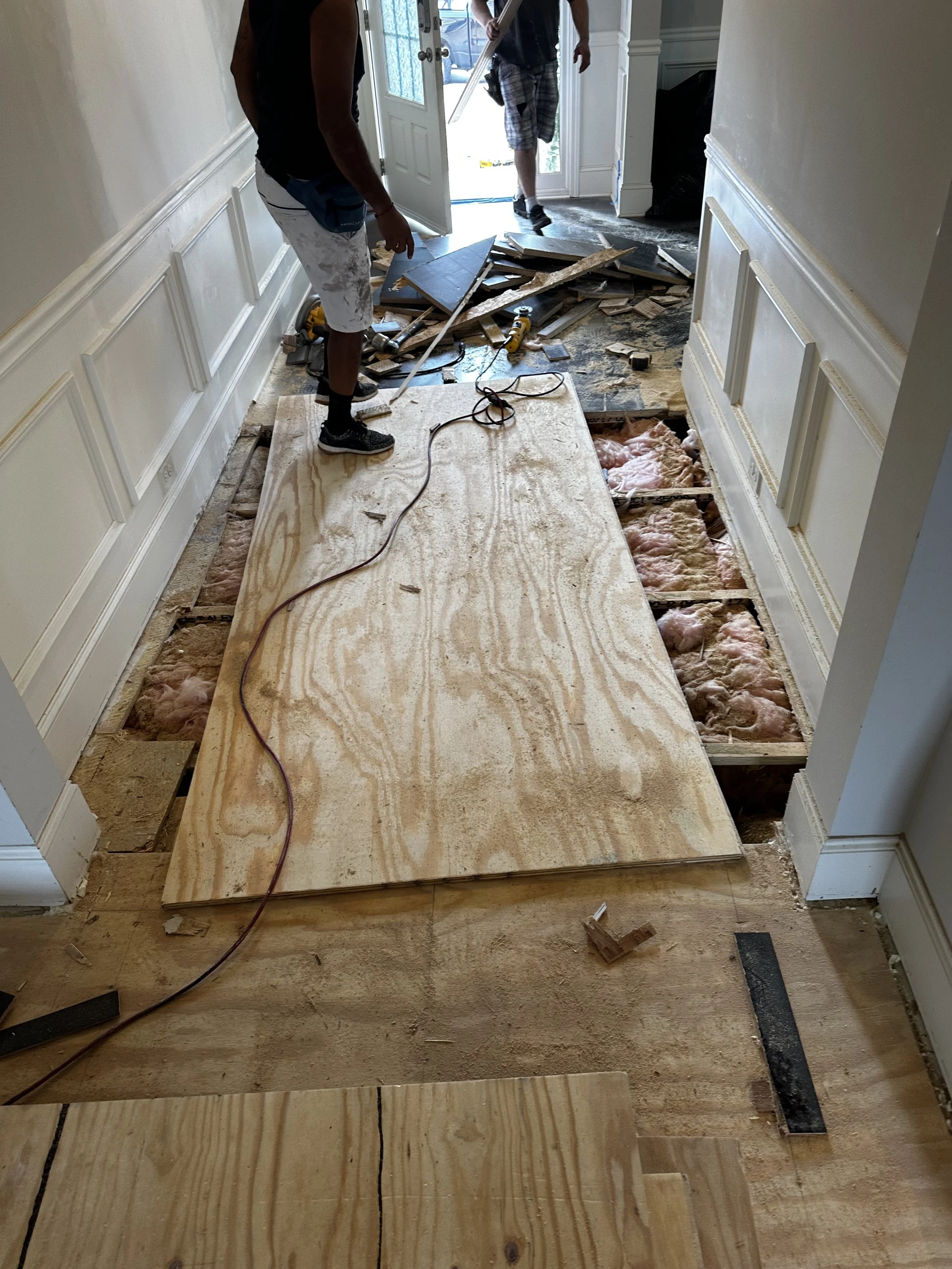 Two men working on a home renovation, removing hardwood flooring. One man stands on a large piece of plywood over cut-out floorboards, using a tool. The other man is in the background near an open door leading outside, holding a piece of flooring.
