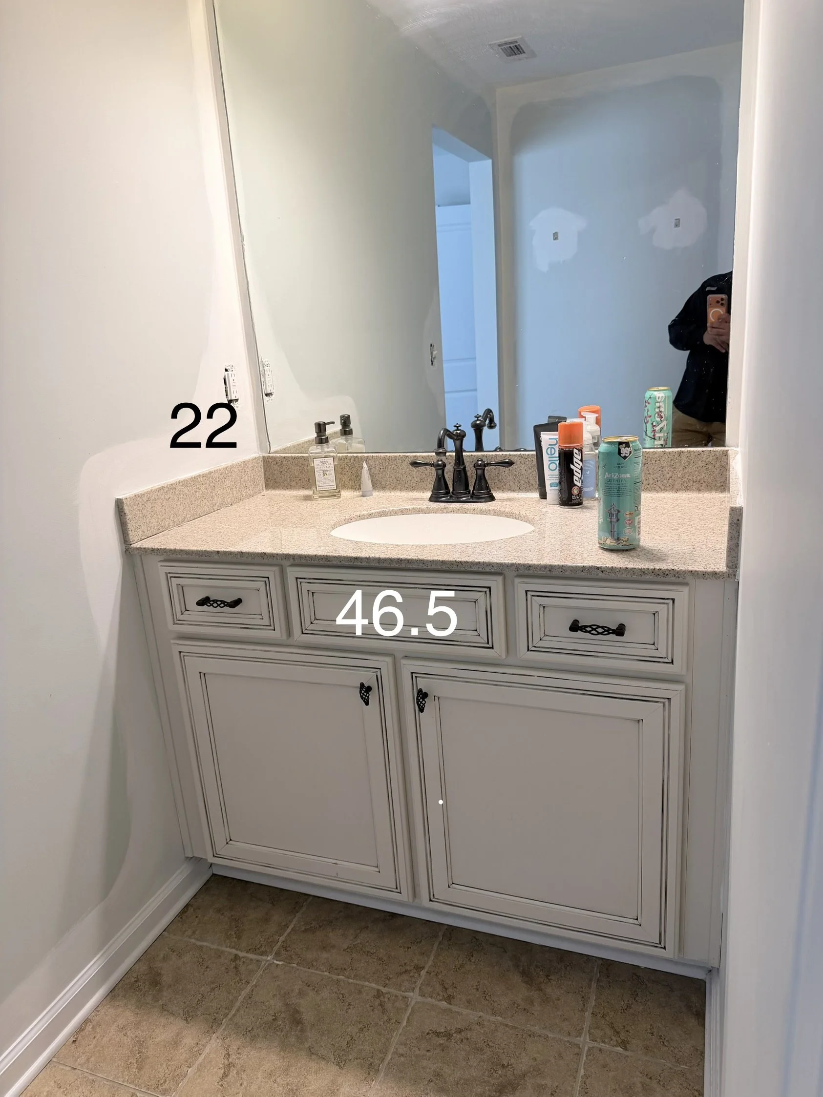 Bathroom vanity with a large mirror, beige countertop, and white cabinetry. Several toiletry bottles and cans are on the counter. A person taking a photo is reflected in the mirror. The wall above the vanity has two patches of drywall. The floor has beige tiles.
