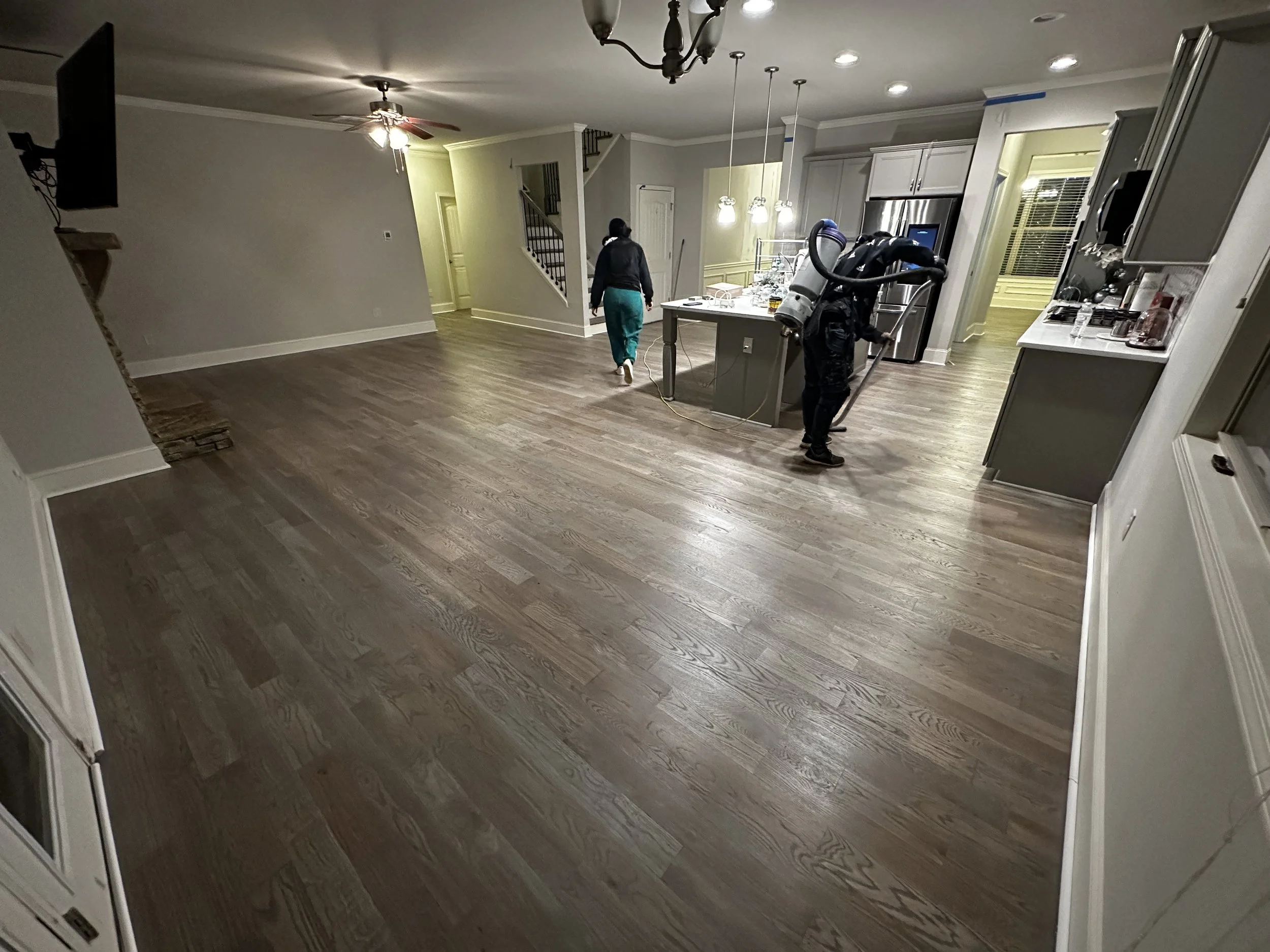 A spacious living room and kitchen area in a house with hardwood floors. Two people are cleaning the floors, one with a vacuum cleaner and the other pushing a cleaning cart, with a ceiling fan and kitchen appliances visible.