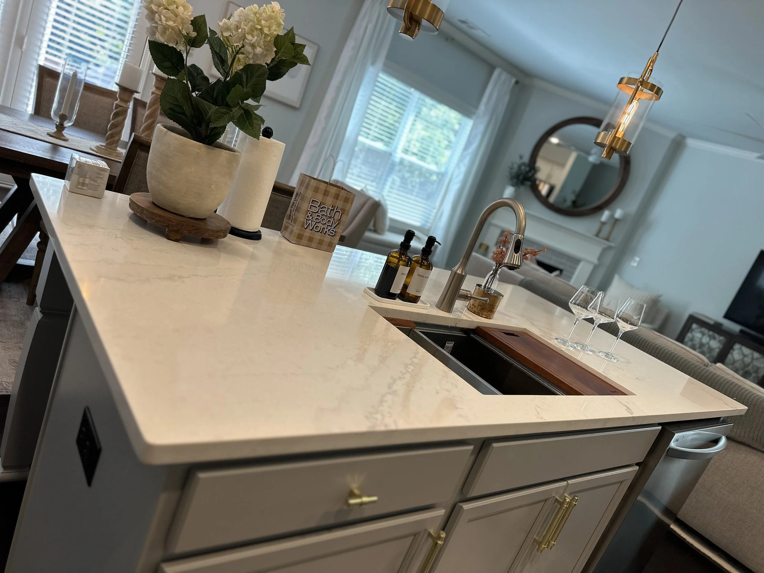 Kitchen island with a white marble countertop, decorative bottles, a potted plant, a paper bag labeled 'Bath & Body Works', and three wine glasses, with a modern sink and brass faucet, in a well-lit open living space.