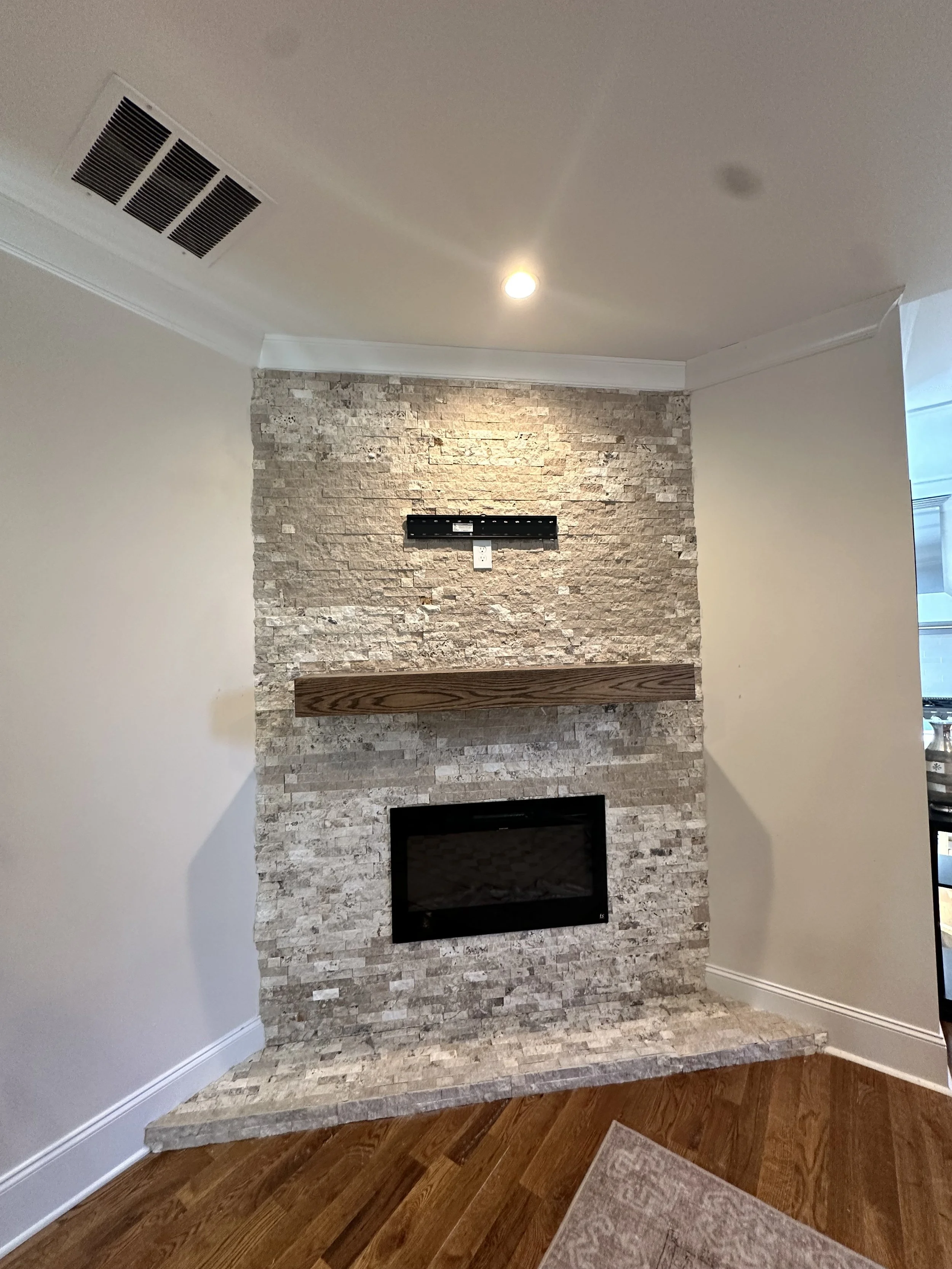 Living room corner with fireplace, stone wall, wooden mantel, TV mount, and ceiling light.