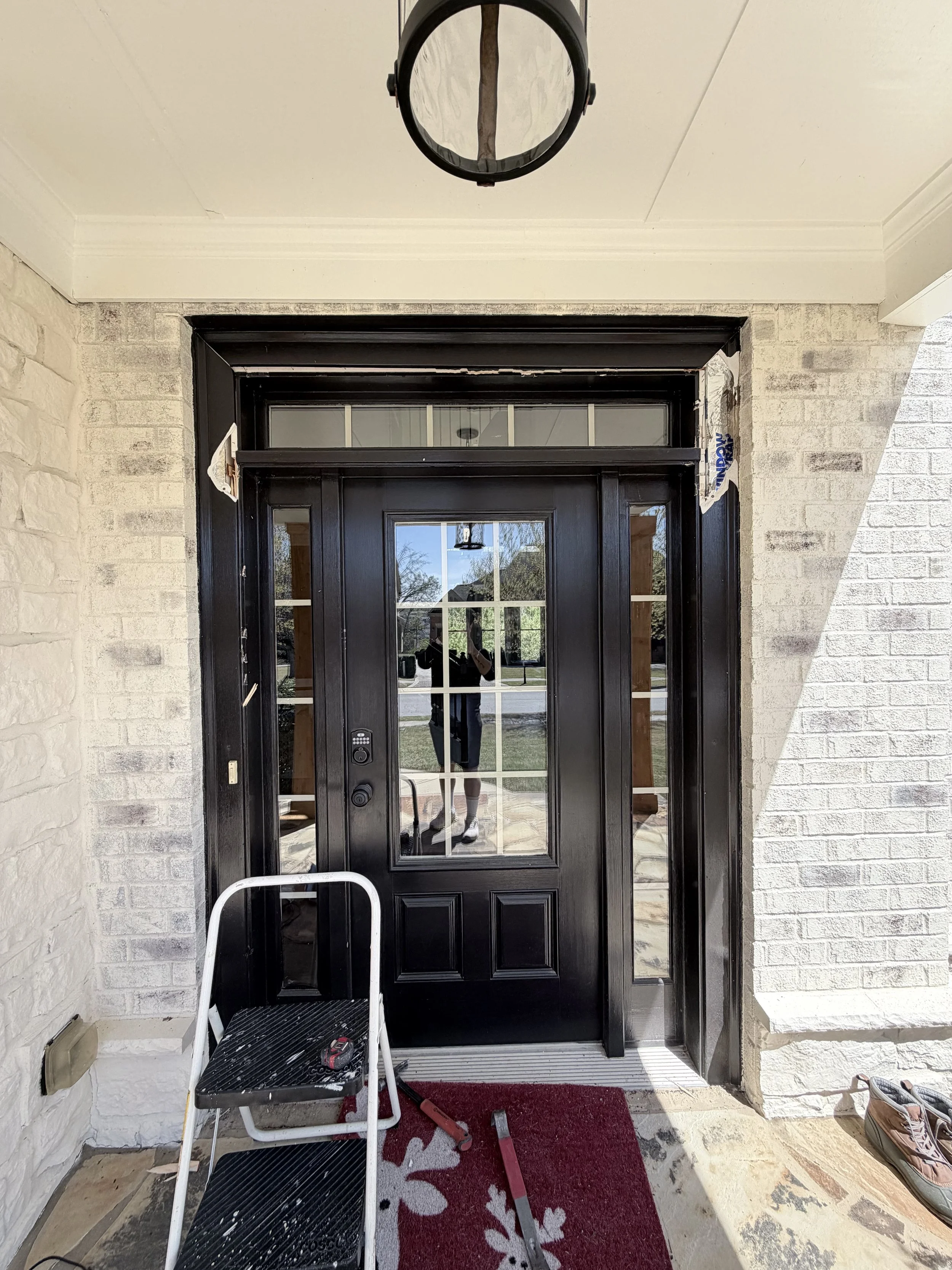 Front entrance door with black frame and glass panels, a step ladder with tools, a red doormat, and shoes to the side, with a partially finished trim around the door.