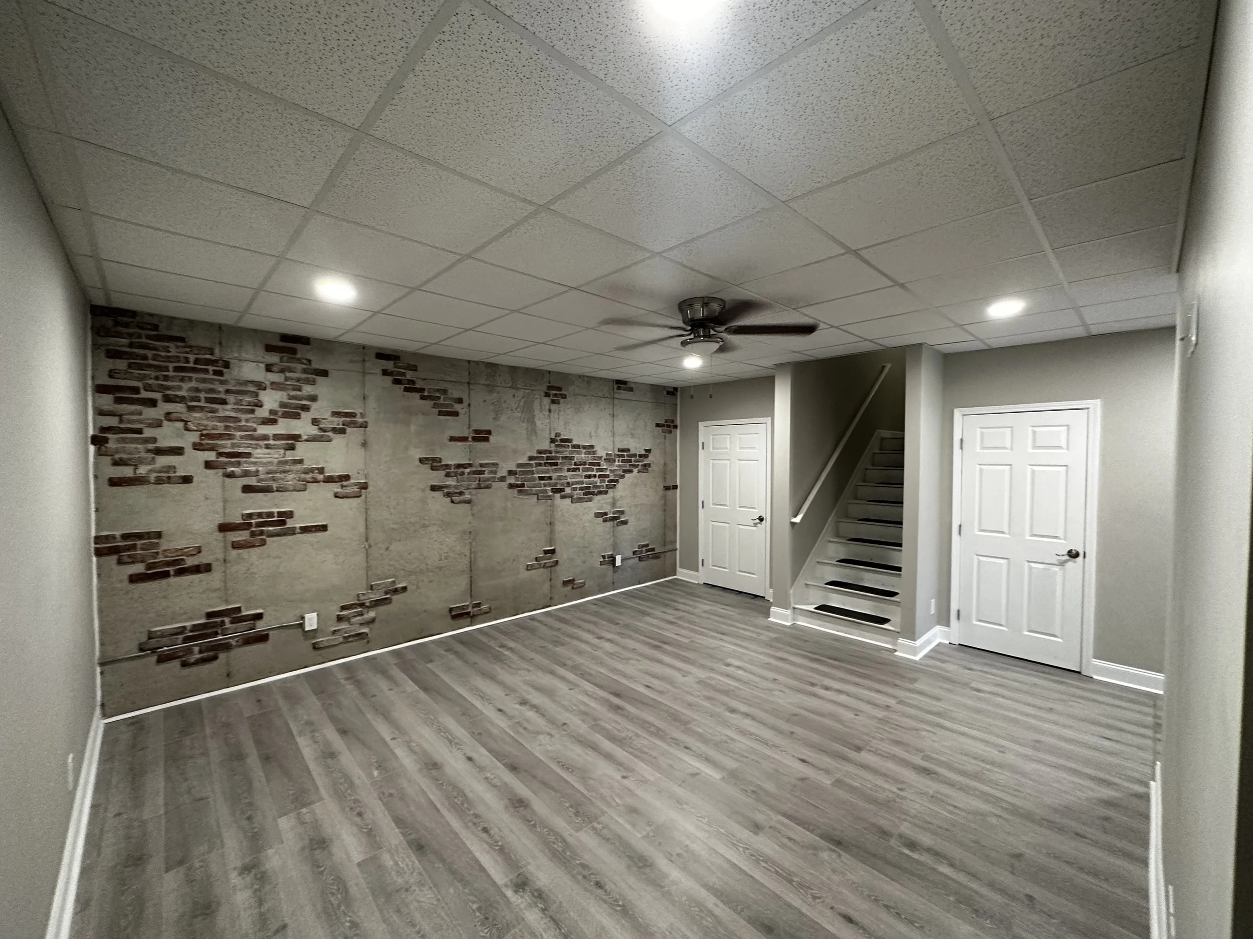 Empty room with a concrete wall with brick accents, light gray walls, gray wood flooring, white doors, ceiling fan, and recessed lighting.