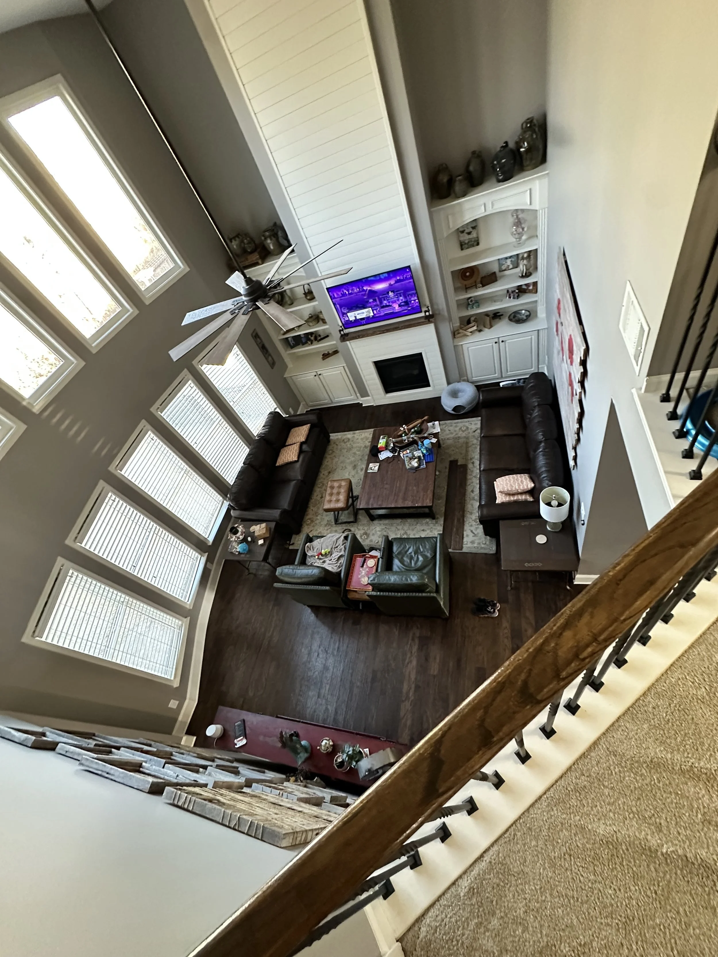 A living room with hardwood flooring, two brown sofas, additional chairs, a coffee table, a white built-in entertainment center with a television, decorative vases, and a wall-mounted artwork. There are large windows allowing natural light, a ceiling fan, and a staircase with a wooden railing.