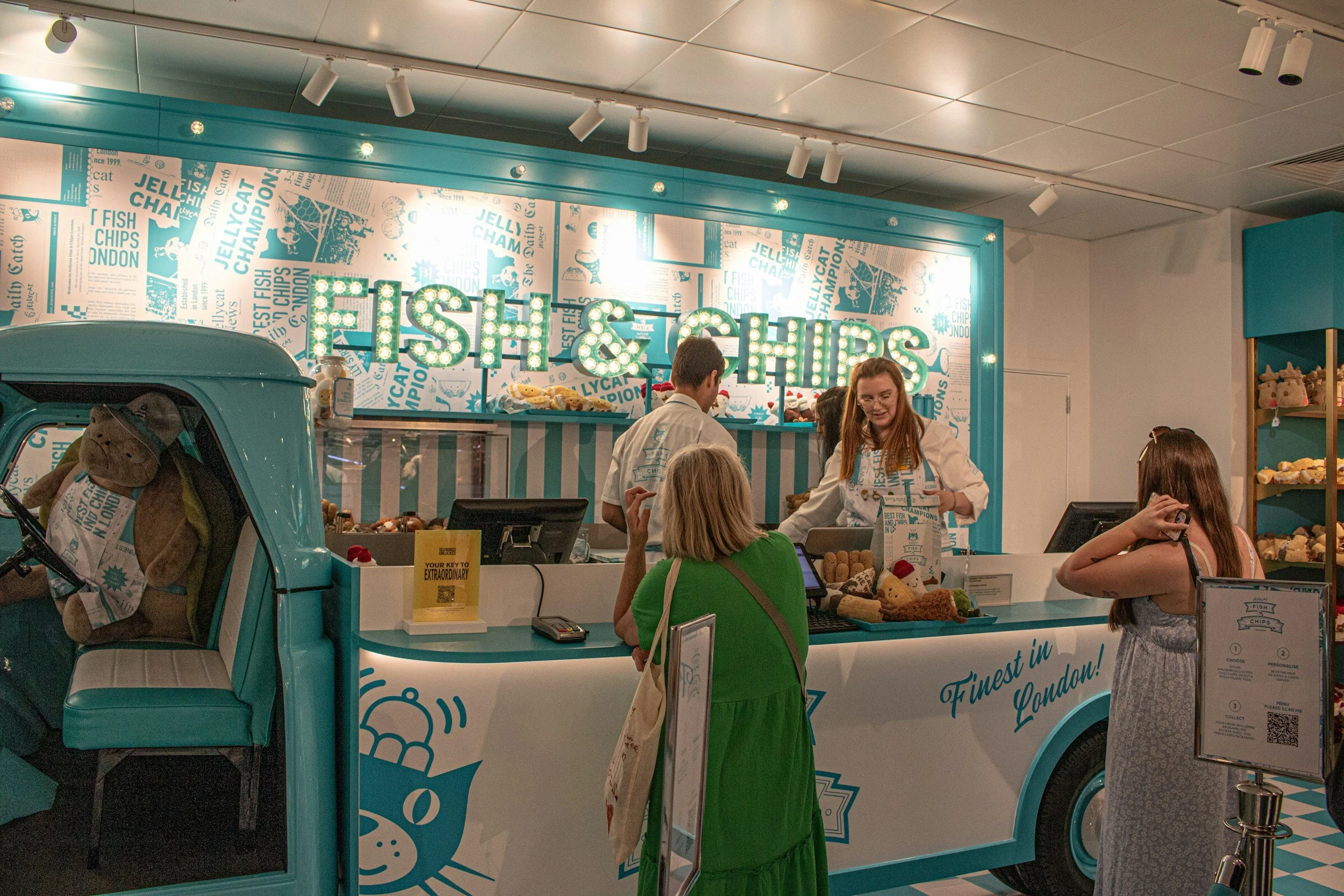 A teal and white food truck labeled 'FISH & CHIPS' with a lit-up sign, inside a mall or food court, with customers ordering at the counter and displays of food, and a shelving unit of stuffed toys in the background.