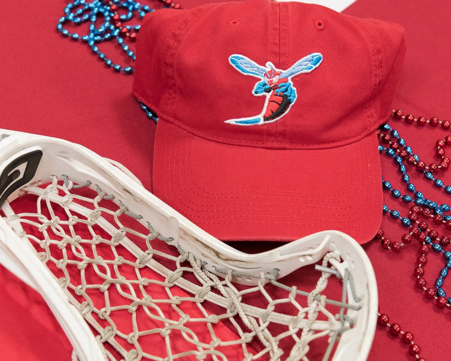 A red baseball cap with a bee logo, a white lacrosse stick, and colorful beads on a red background.