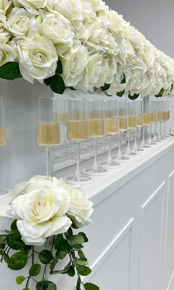 Elegant arrangement of white roses and hydrangeas with champagne glasses on a white countertop.