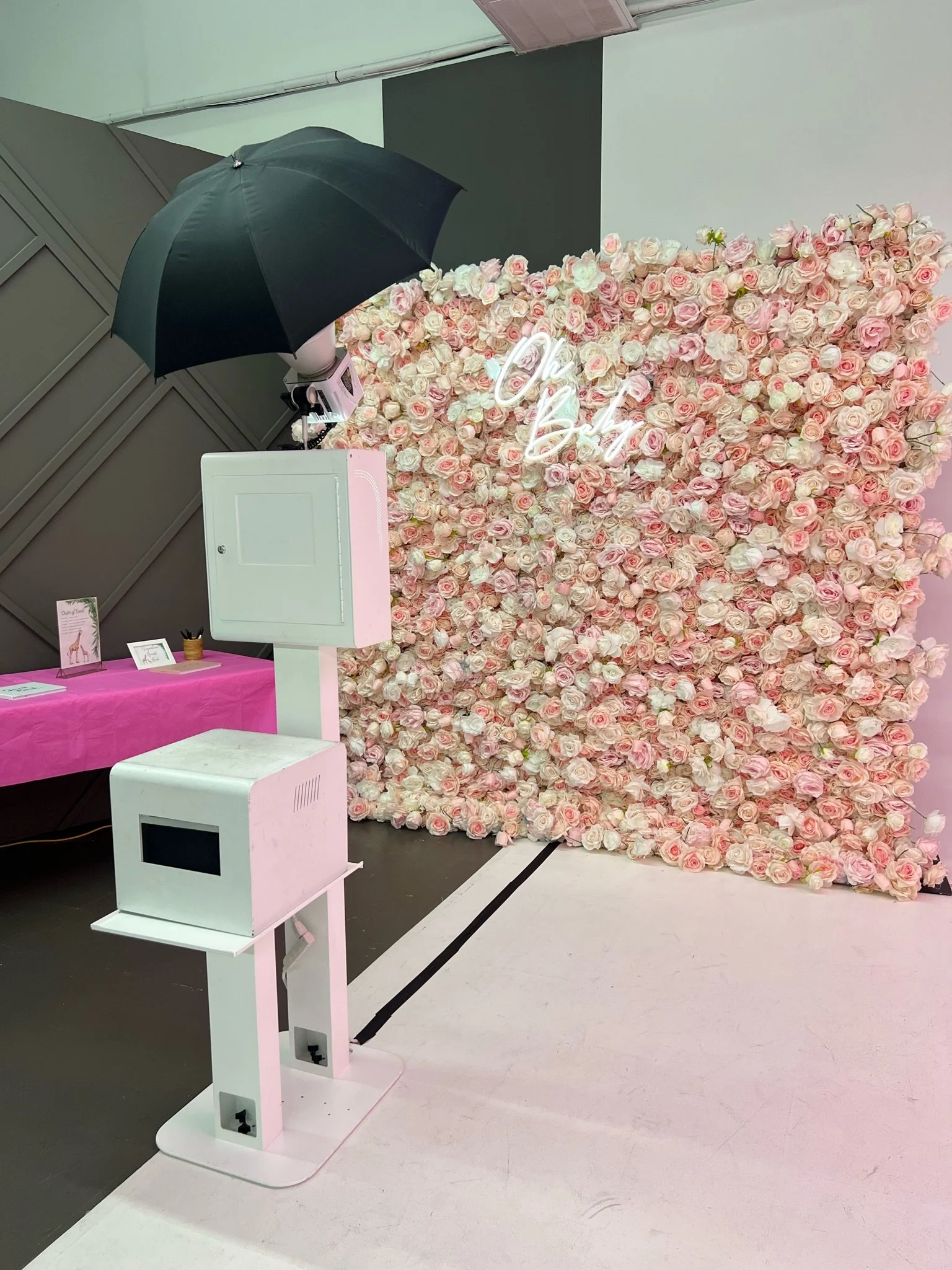 A photo booth setup with a backdrop of pink and white roses, neon sign that says 'Oh, Baby,' a black umbrella for lighting, and photo booth equipment including a camera and printing station. There is a table with pink tablecloth and decorative items 