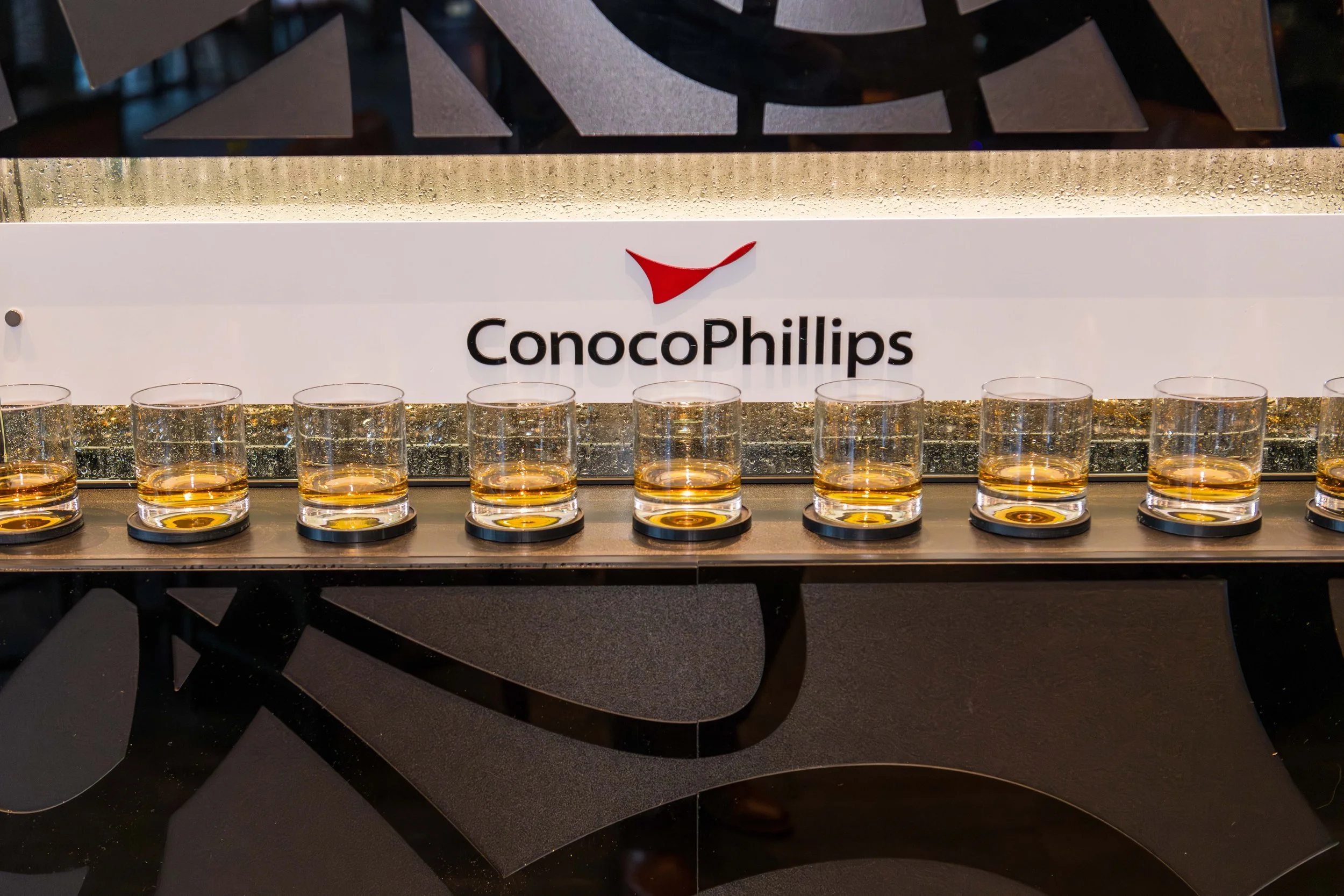 Eight glasses of whiskey on a black counter with a ConocoPhillips sign in the background.