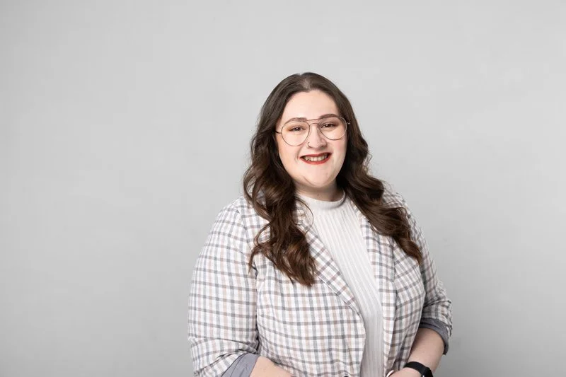 A young woman with long dark brown hair, wearing glasses, a checkered blazer, and a white top, smiling at the camera against a light gray background.