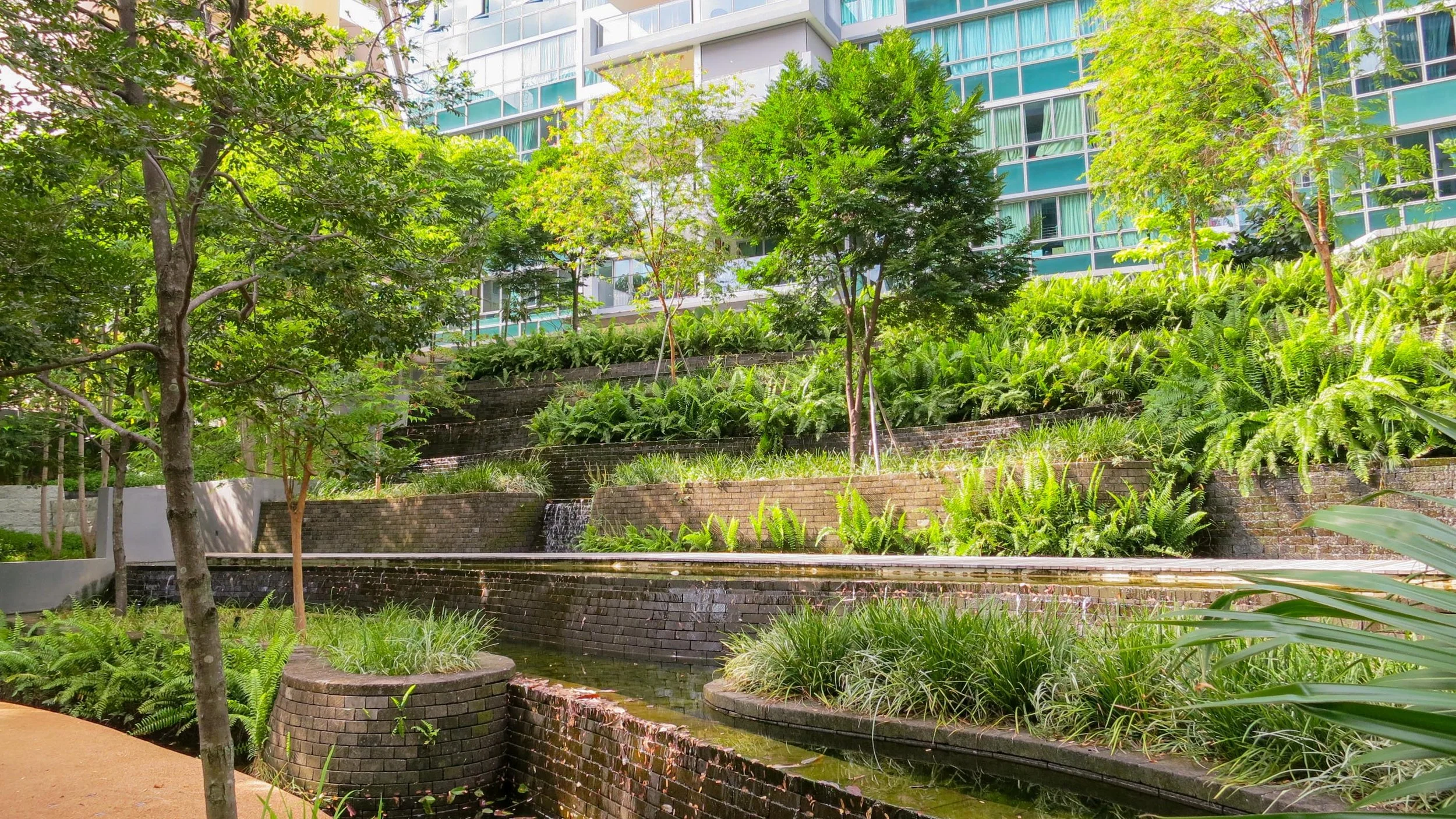 A modern urban park with multiple tiered water features, lush greenery, and trees, set against a backdrop of a high-rise glass building.