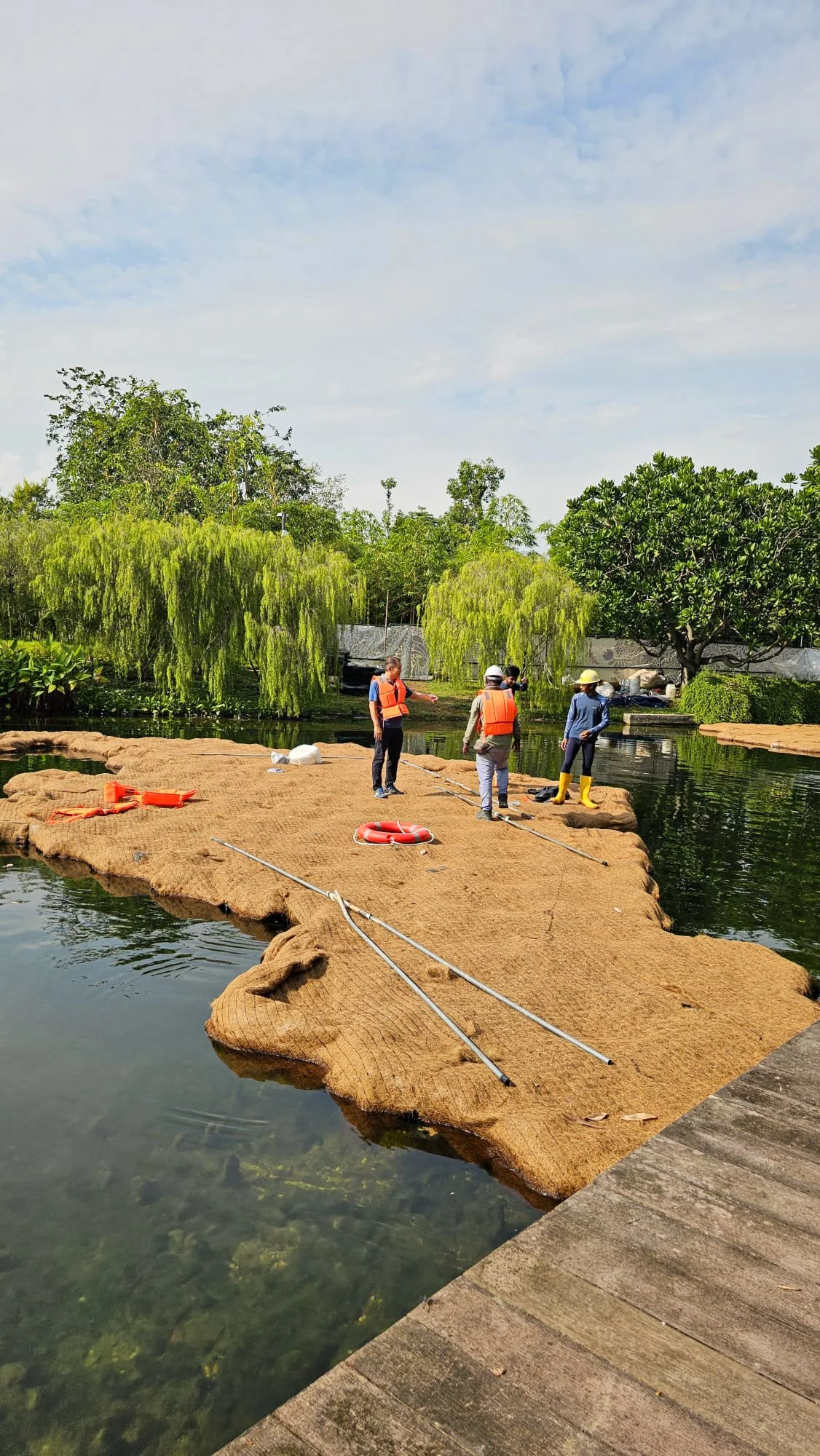 Four people in life vests and hats standing on a floating platform on a river, surrounded by trees with green leaves, with life-saving equipment nearby.