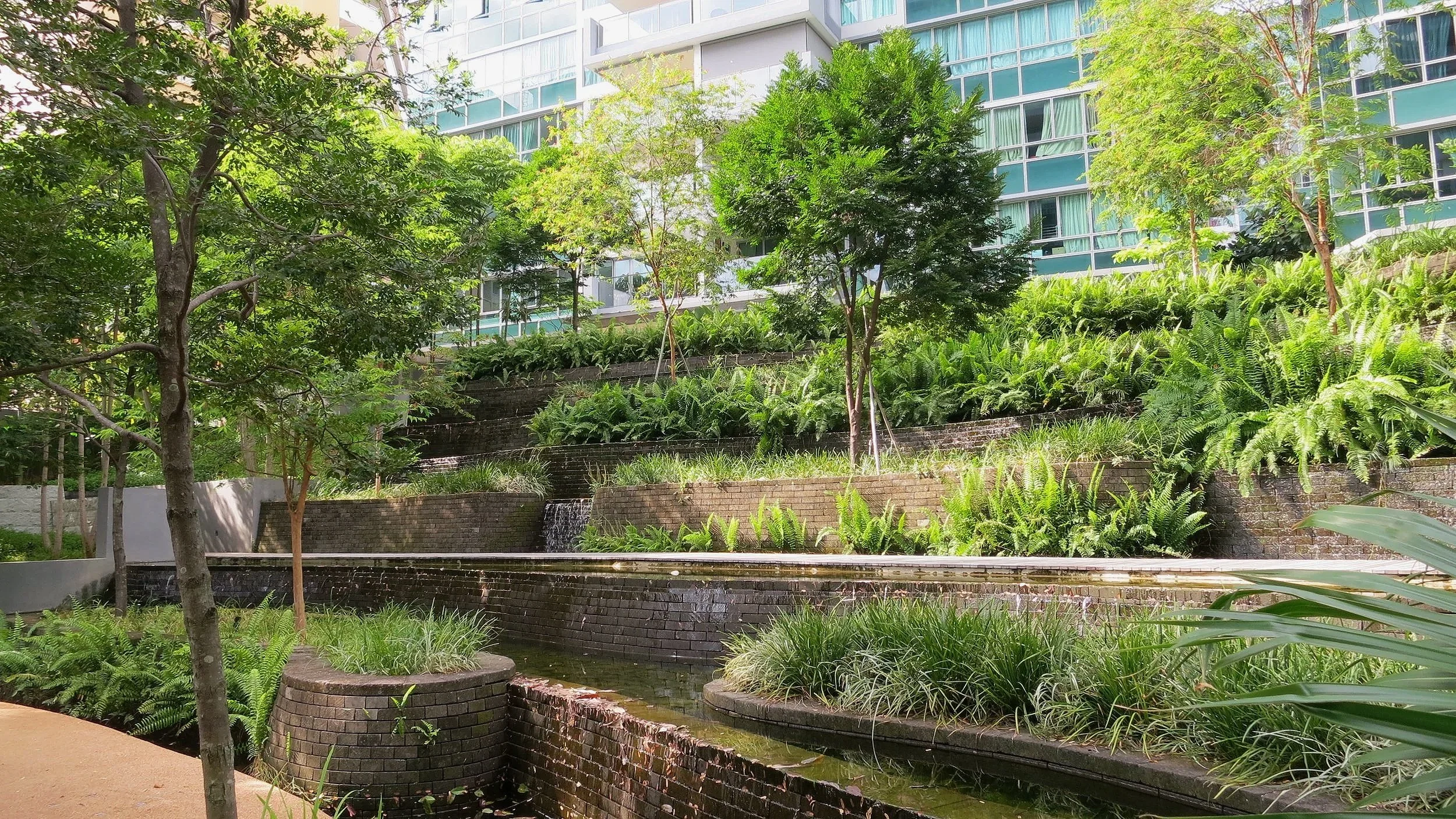 A landscaped urban garden with tiered brick retaining walls, lush green plants, small trees, and a modern glass building in the background.