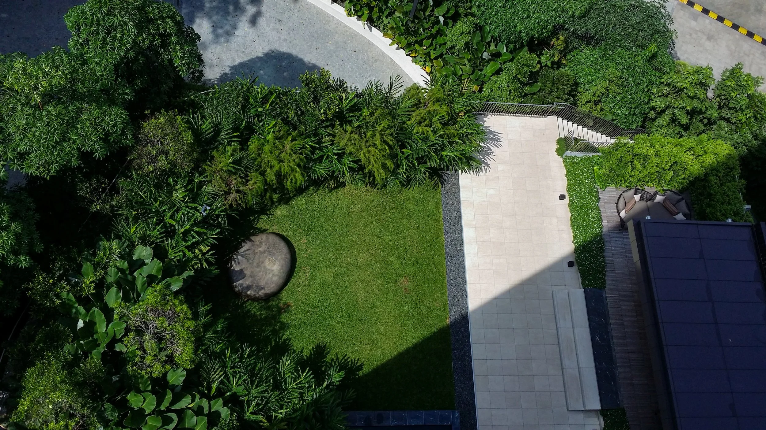 An aerial view of a modern backyard with lush green trees, a well-maintained lawn, a paved patio area, and outdoor furniture, surrounded by a privacy fence.