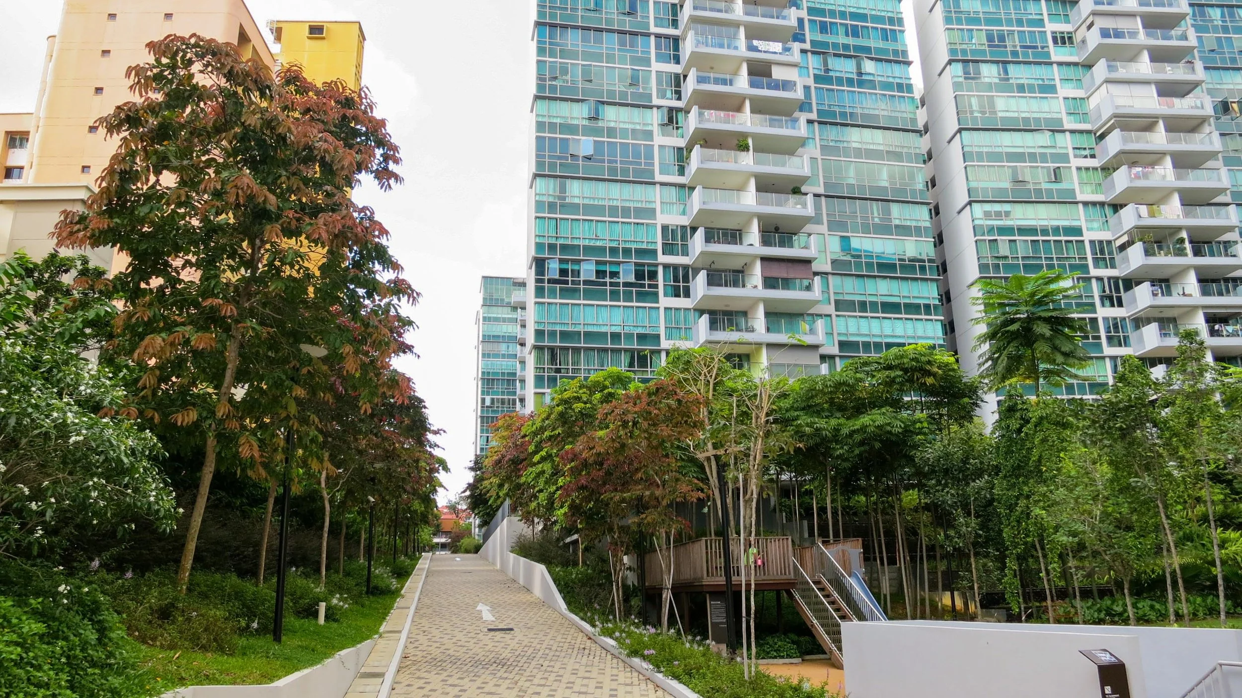 Tall modern apartment building with glass balconies and a lush green park with trees and a paved pathway in the foreground.