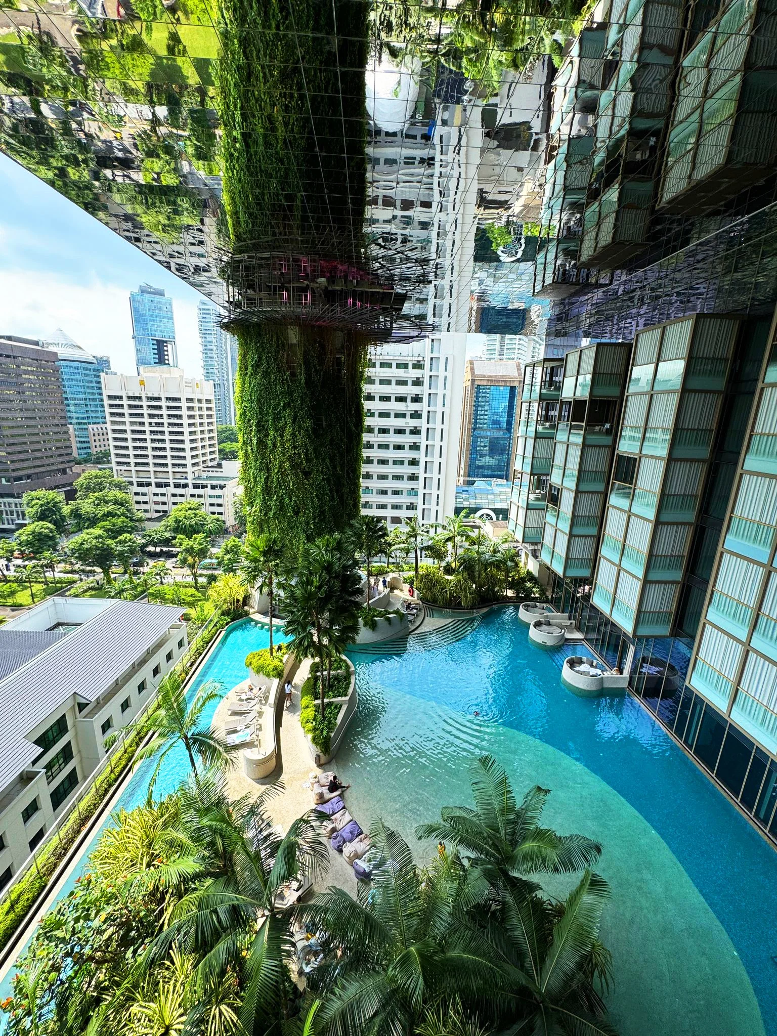 View of a cityscape with tall buildings, a swimming pool, lounge chairs, and palm trees, taken from a high balcony or rooftop on a sunny day.