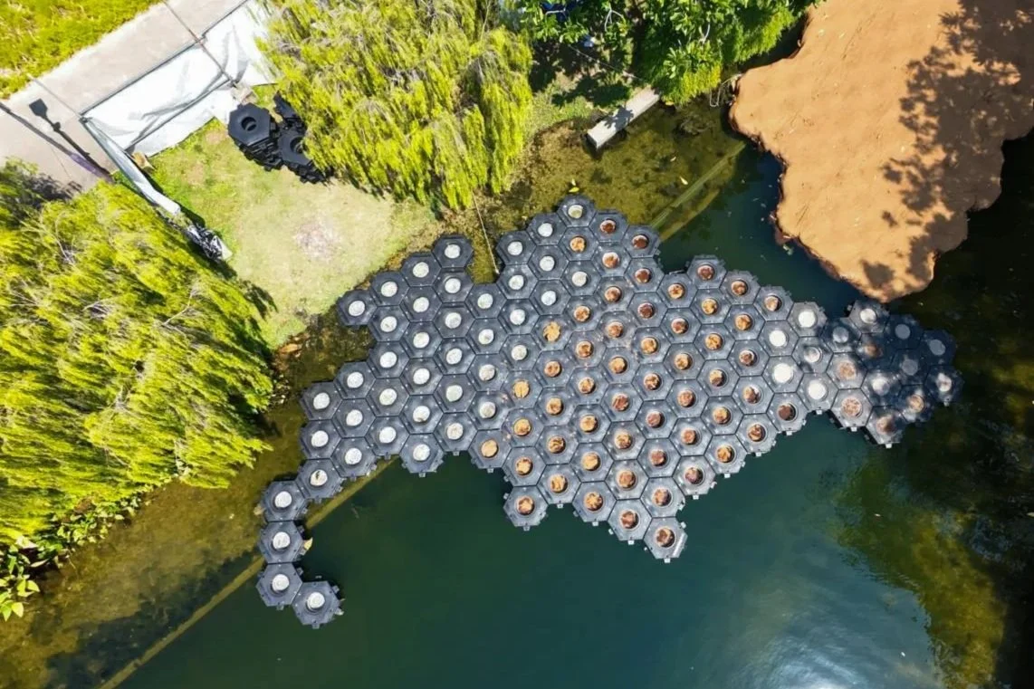 Drone view of a floating pond with hexagonal black and white floating platforms, forming a shape of the state of Texas, surrounded by green trees and a body of water.