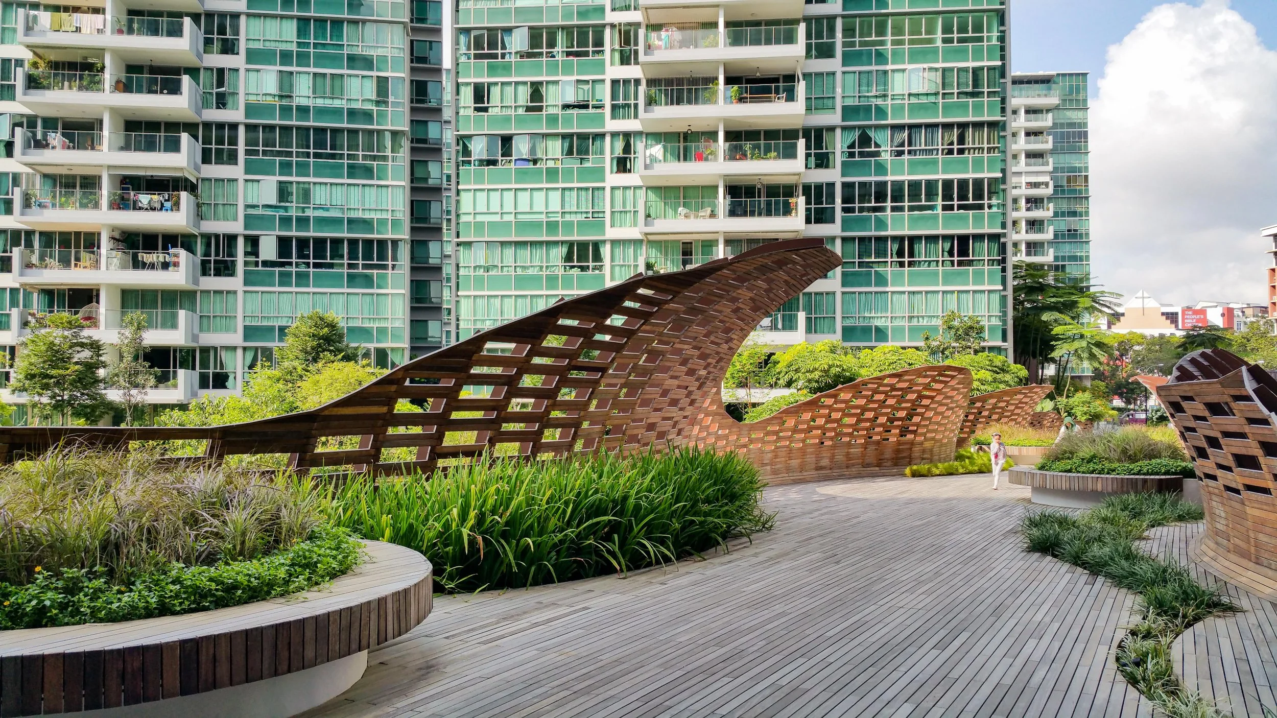 Modern high-rise apartment building with glass balconies overlooking a landscaped park with wooden benches, trees, and walking paths.