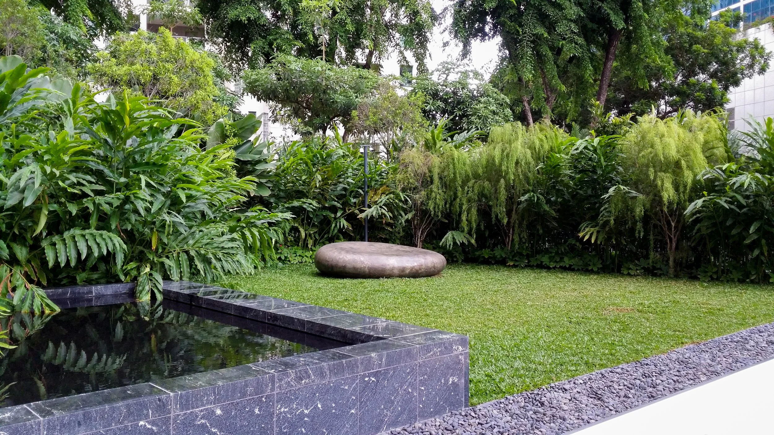 Urban garden with lush green trees and bushes, a stone bench, a small waterfall, and a gravel pathway in front, surrounded by tall buildings.