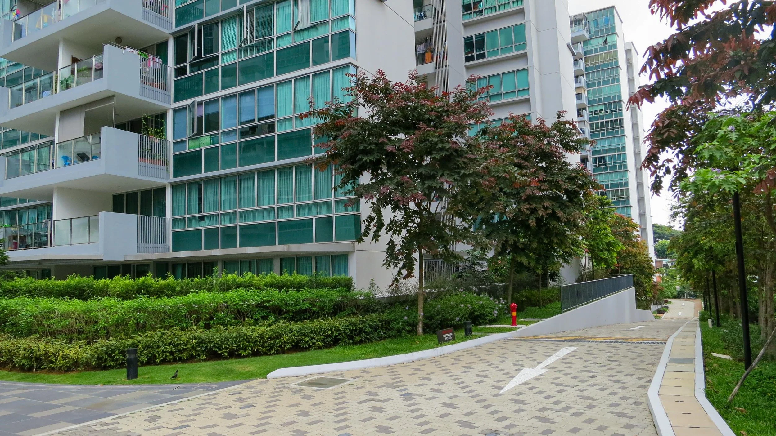 View of a modern apartment building with glass windows and balconies, surrounded by trees and greenery, with a paved walking path in the foreground.