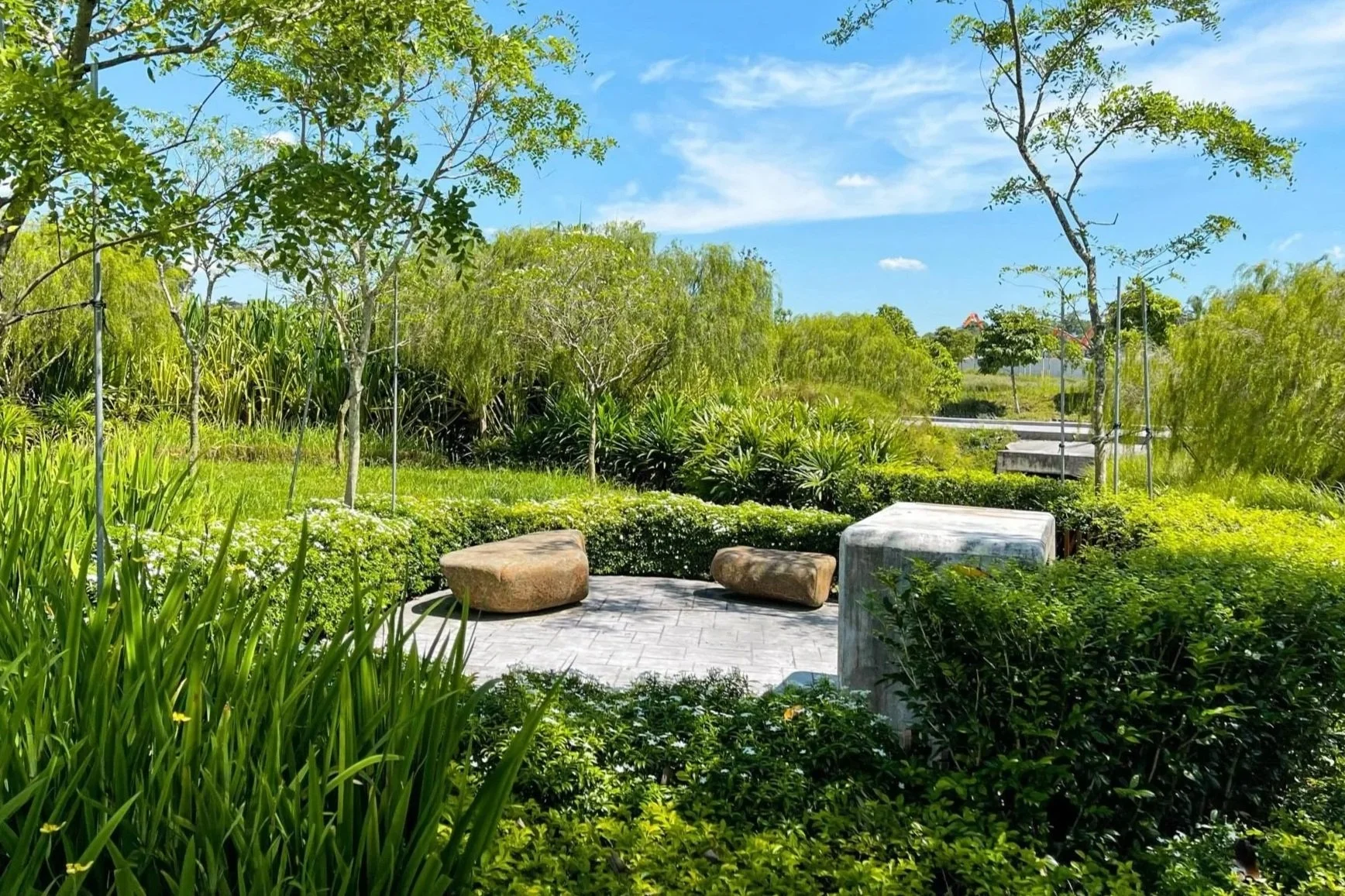 A serene outdoor scene of a park with greenery including trees, bushes, and grass, with a small paved area featuring large rocks and a marble bench, under a clear blue sky with some clouds.