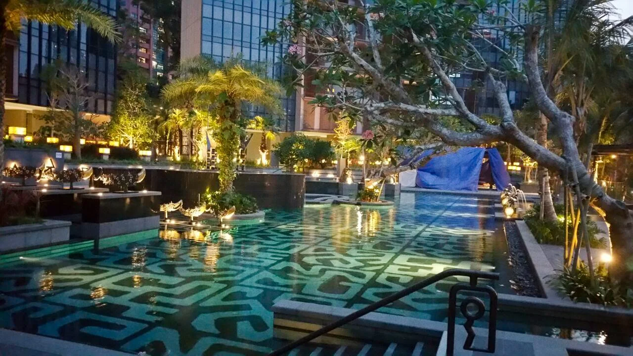 Luxury rooftop pool area at dusk with lit planters, trees, and city buildings in the background.