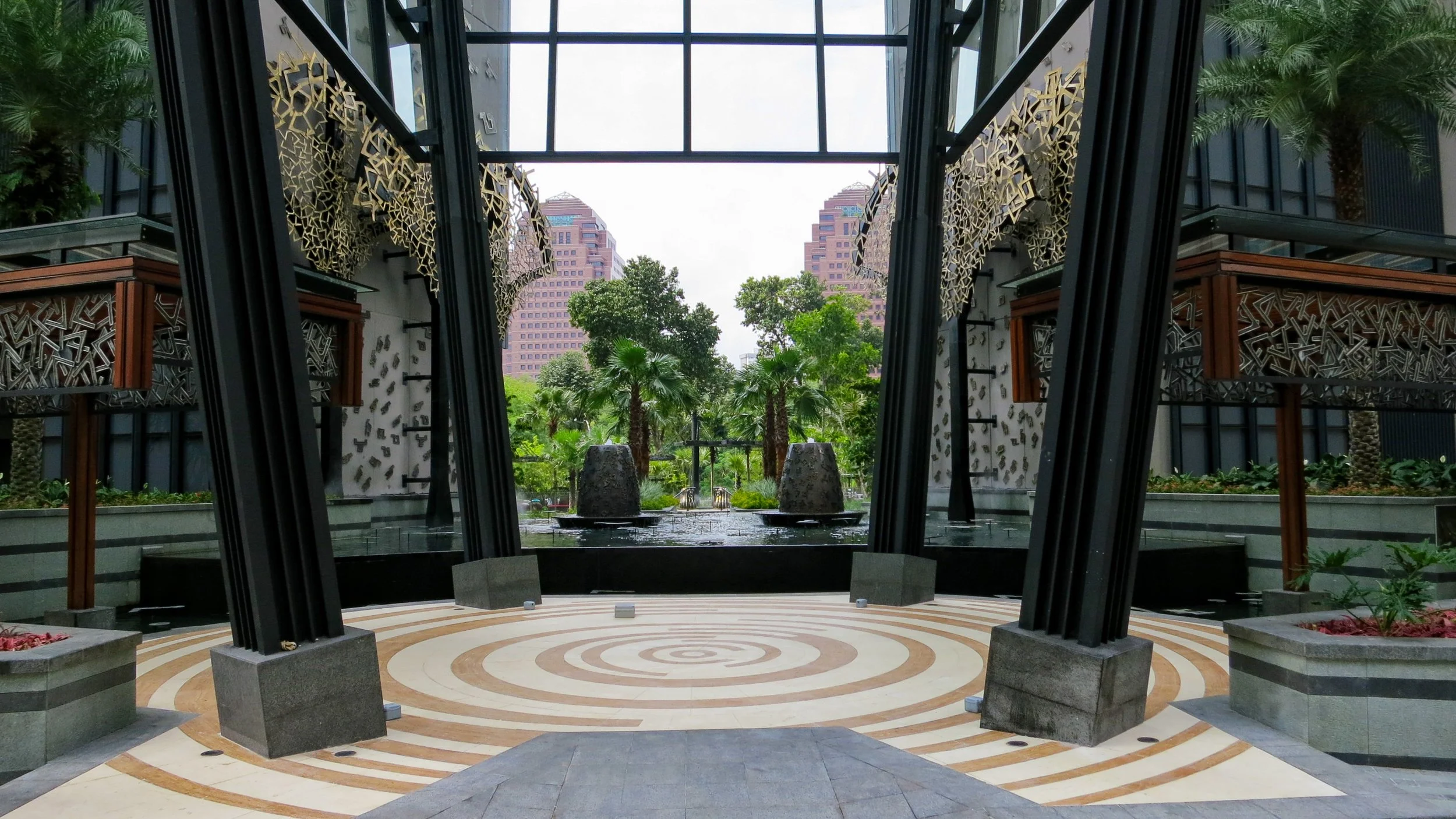 Indoor area with modern architectural design, featuring black structural supports, decorative gold elements on the walls, a patterned circular floor, and a view of a lush garden with trees through a glass ceiling.