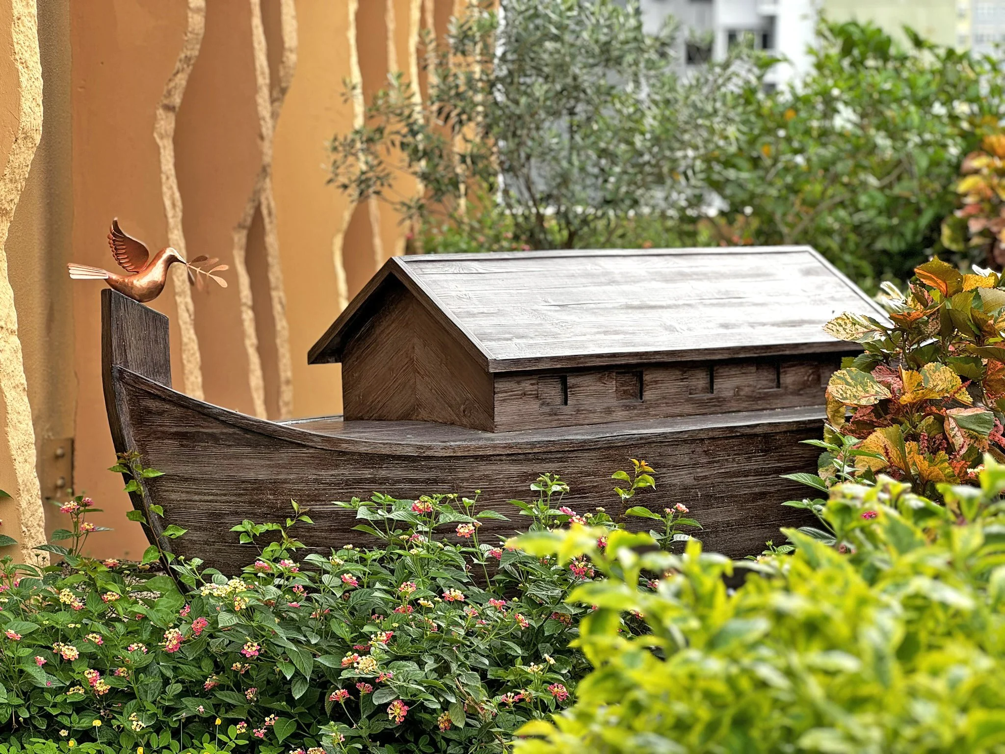 A decorative metal bird sculpture perched on the edge of a wooden garden bed, surrounded by green and yellow plants and flowers, with a brick wall on the left and blurred trees and buildings in the background.